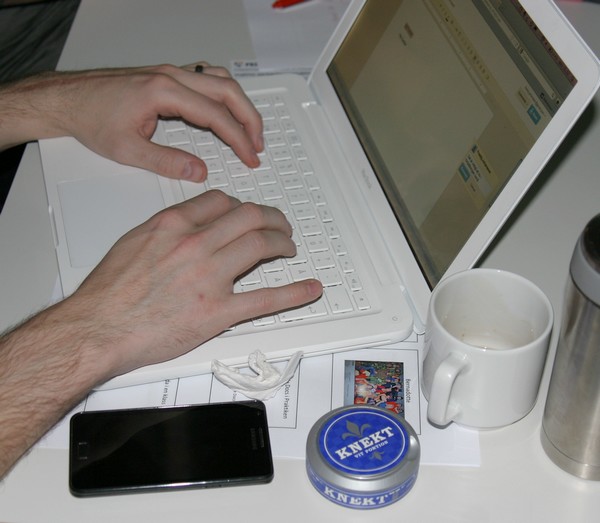 Händer skriver på en vit macbook, vid sidan ligger mobiltelefon, kaffekopp och en snusdosa.