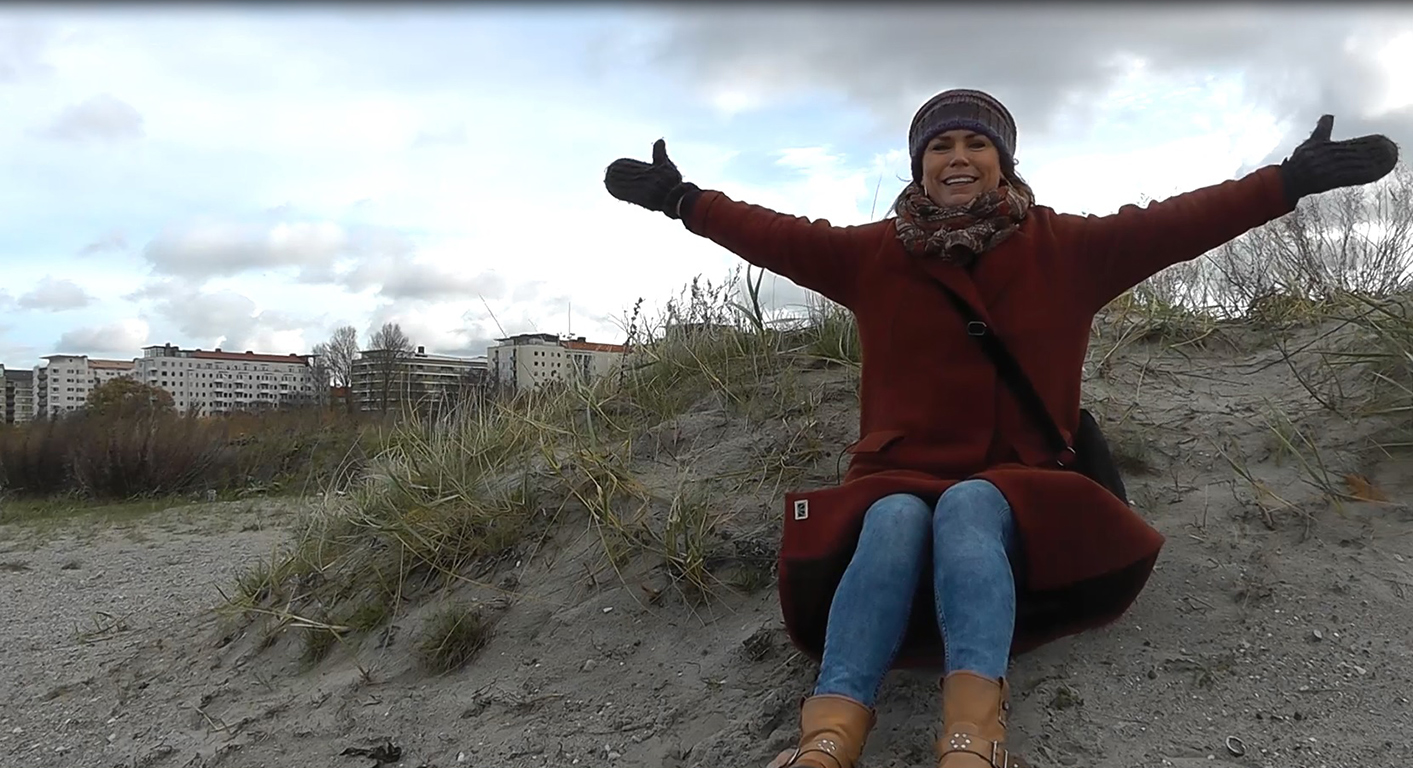 Kvinna sitter på strand och slår ut med armarna.