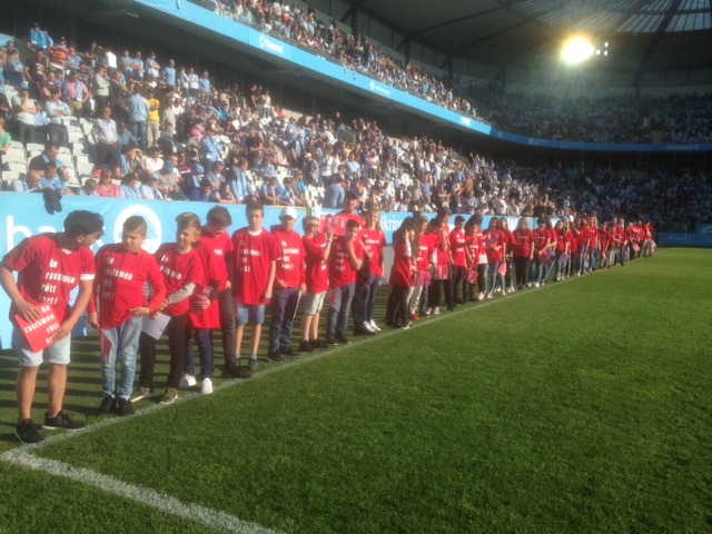 Elever i röda t-shirtar står vid linjen av fotbollsplan.