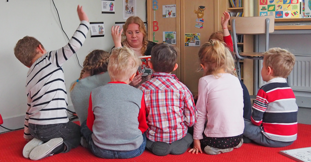 Kvinna läser bok framför barn som sitter på golvet.