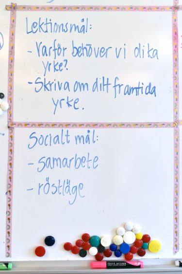 Lektionsmål och sociala mål står på whiteboard. Lektionsmål: Varför behöver vi olika yrken, Skriva om ditt framtida yrke. Socialt mål: Samarbete, röstläge.