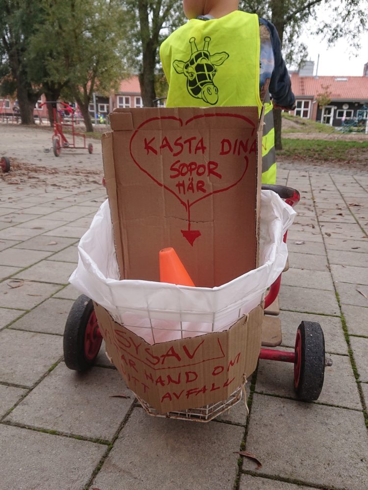 Barn cyklar på en trehjuling omgjord till sopbil. "Kasta dina sopor här" står det på den.