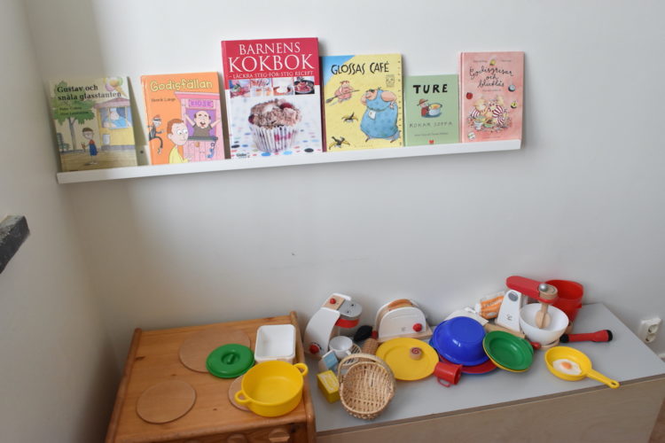 Kokböcker för barn uppställda på hylla. Under den köksredskapsleksaker.