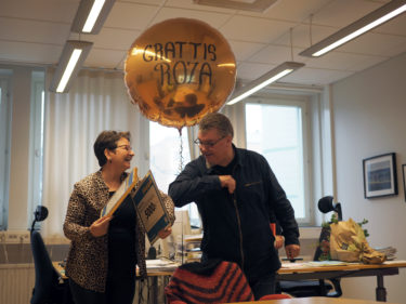 Roza och Anders nuddar armbågar mot varandra och i bakgrunden svävar en stor ballong där det står grattis Roza!