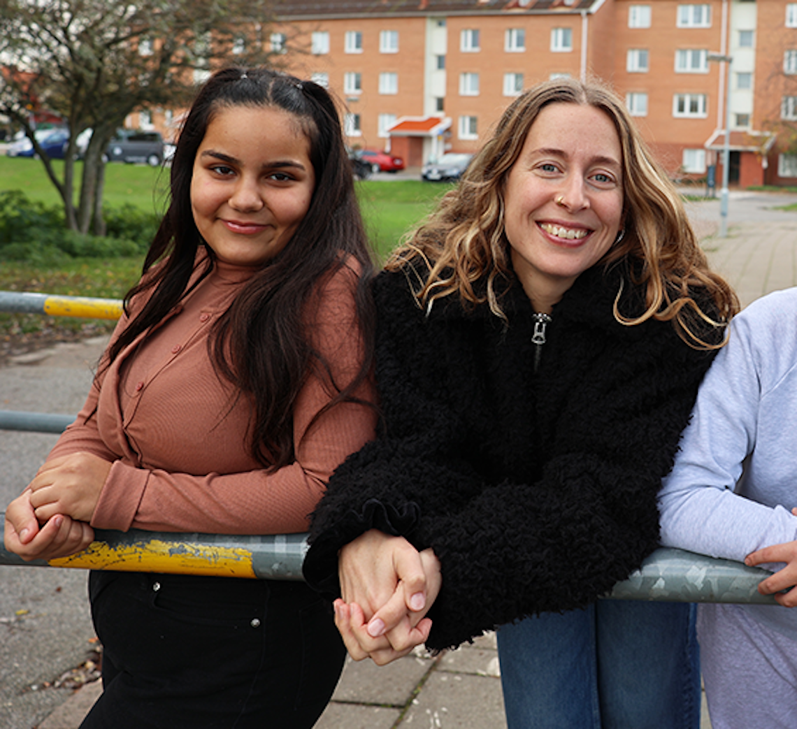 Två personer står lutade mot ett räcke på en gård med bostadshus i bakgrunden.