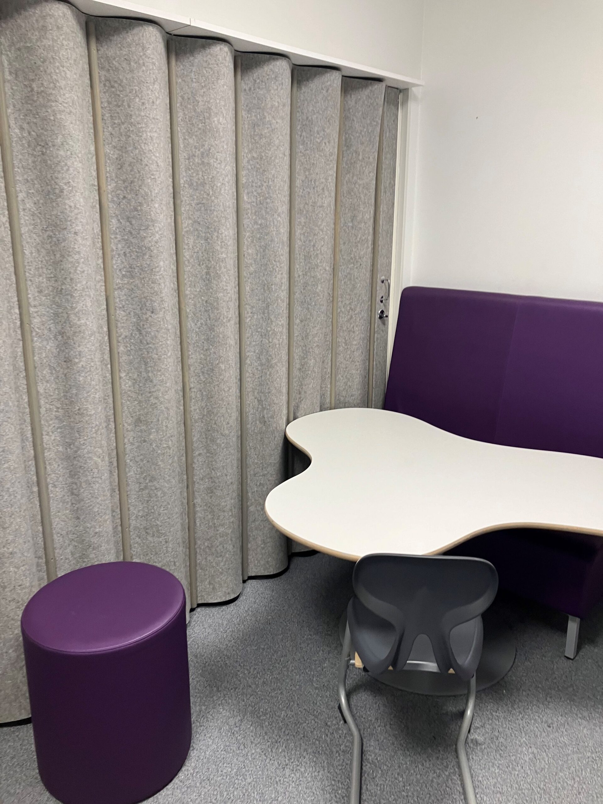 Utifrån väggen finns en vikbar vägg, intill väggen finns ett bord i organisk form och en soffa intill väggen. På andra sidan bordet står en cylinderformad pall.