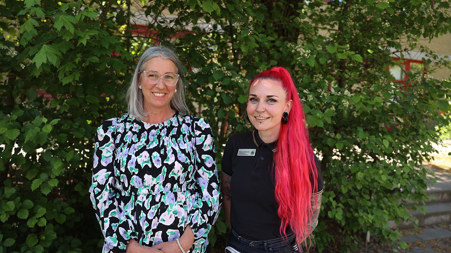 Två personer står utomhus framför en grön buske.