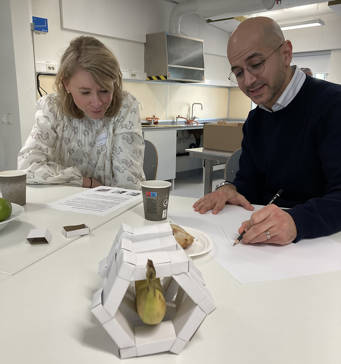 Arkitekterna Haydar Alward och Kristina Kember sitter vid ett bord och ritar. I förgrunden står en modell av vita tändsticksaskar som omsluter en banan.