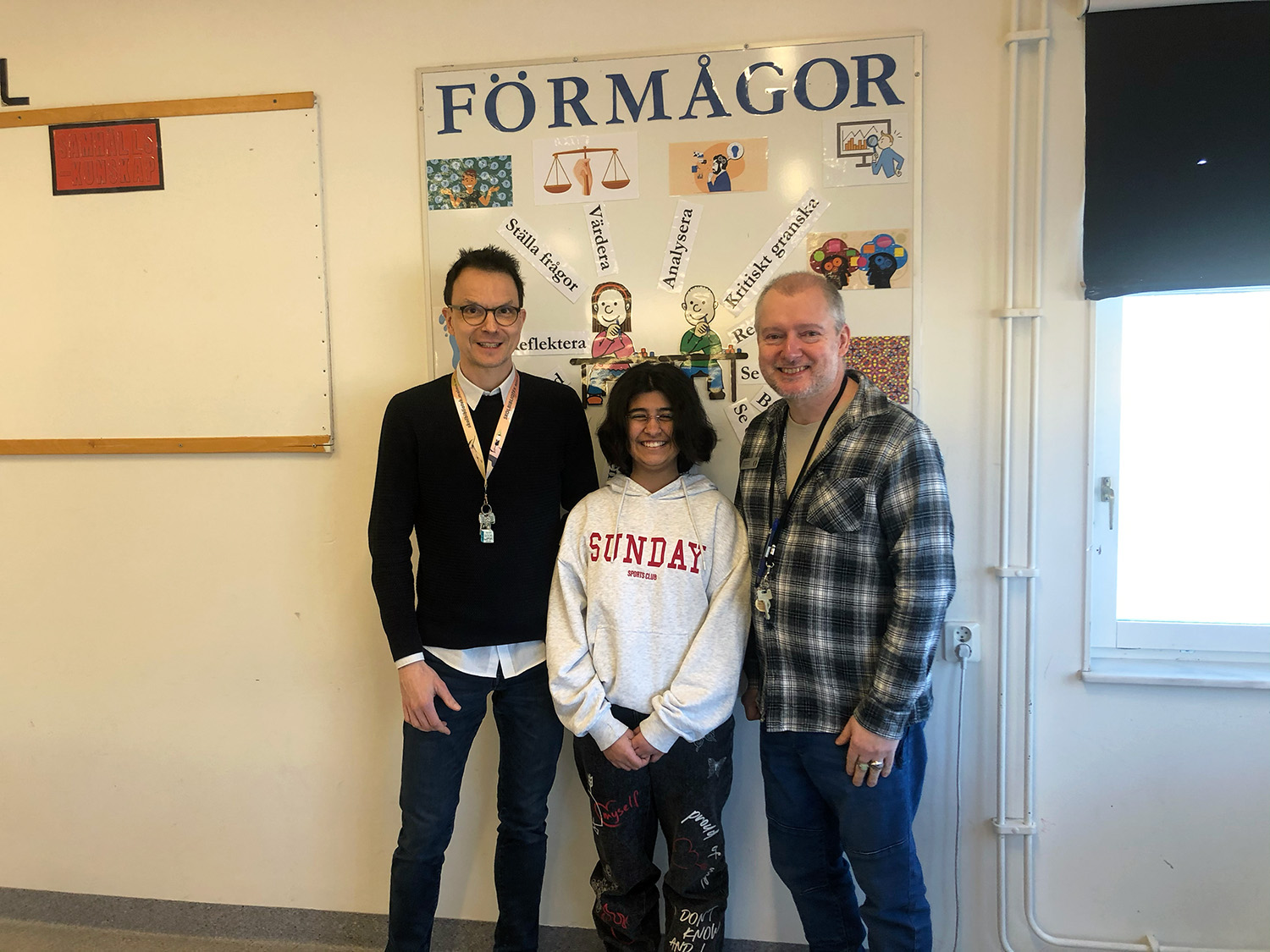 Två lärare och en elev poserar framför ett collage med rubriken 'Förmågor'. Där listas bland annat 'värdera', 'analysera' och 'kritiskt tänkande'.