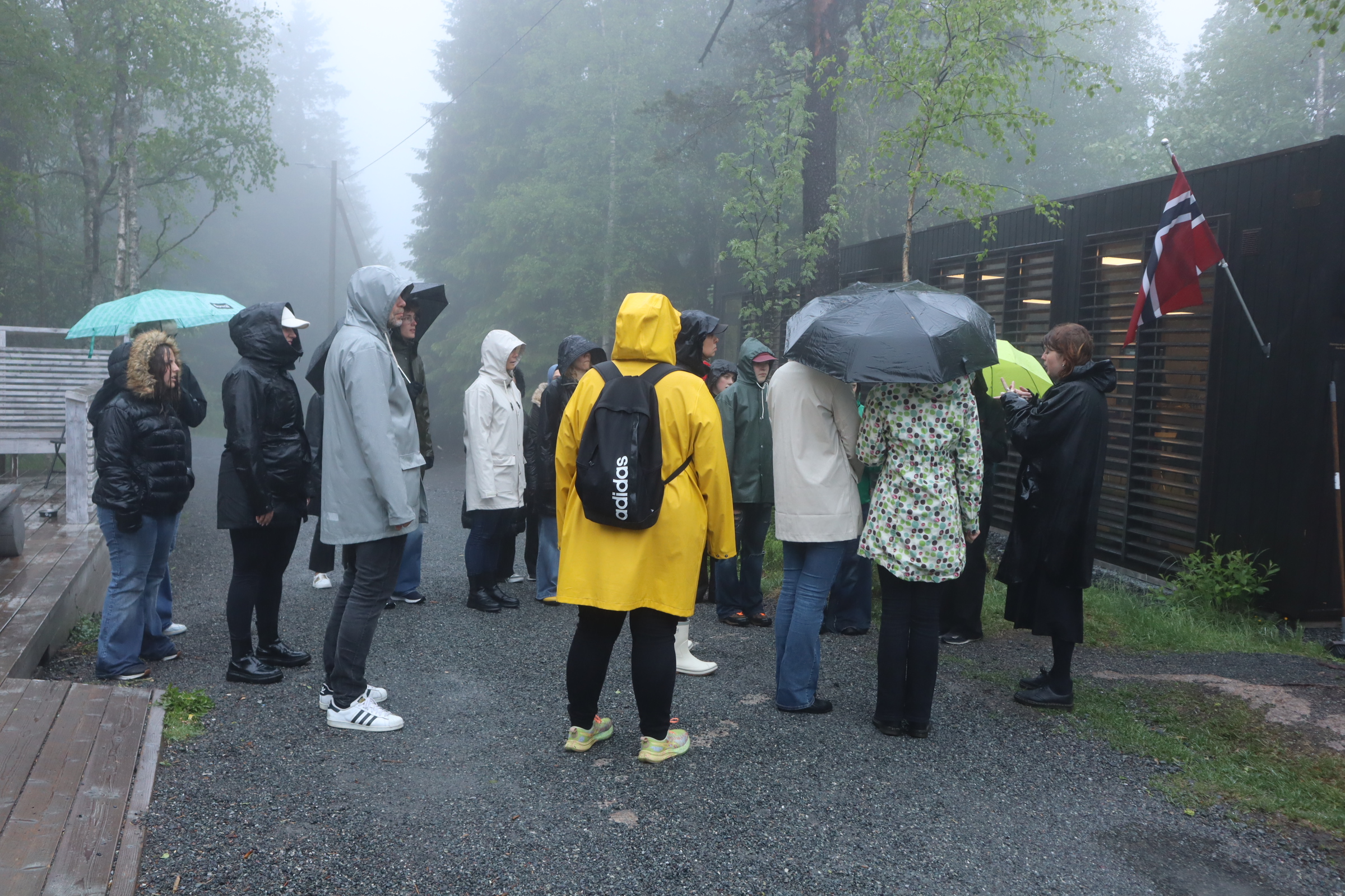 En samling elever och lärare står i regnkläder vid en norsk flagga.