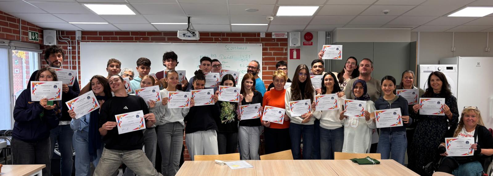 En grupp personer står i ett klassrum och håller upp diplom, framför en whiteboard med text och en tegelvägg.