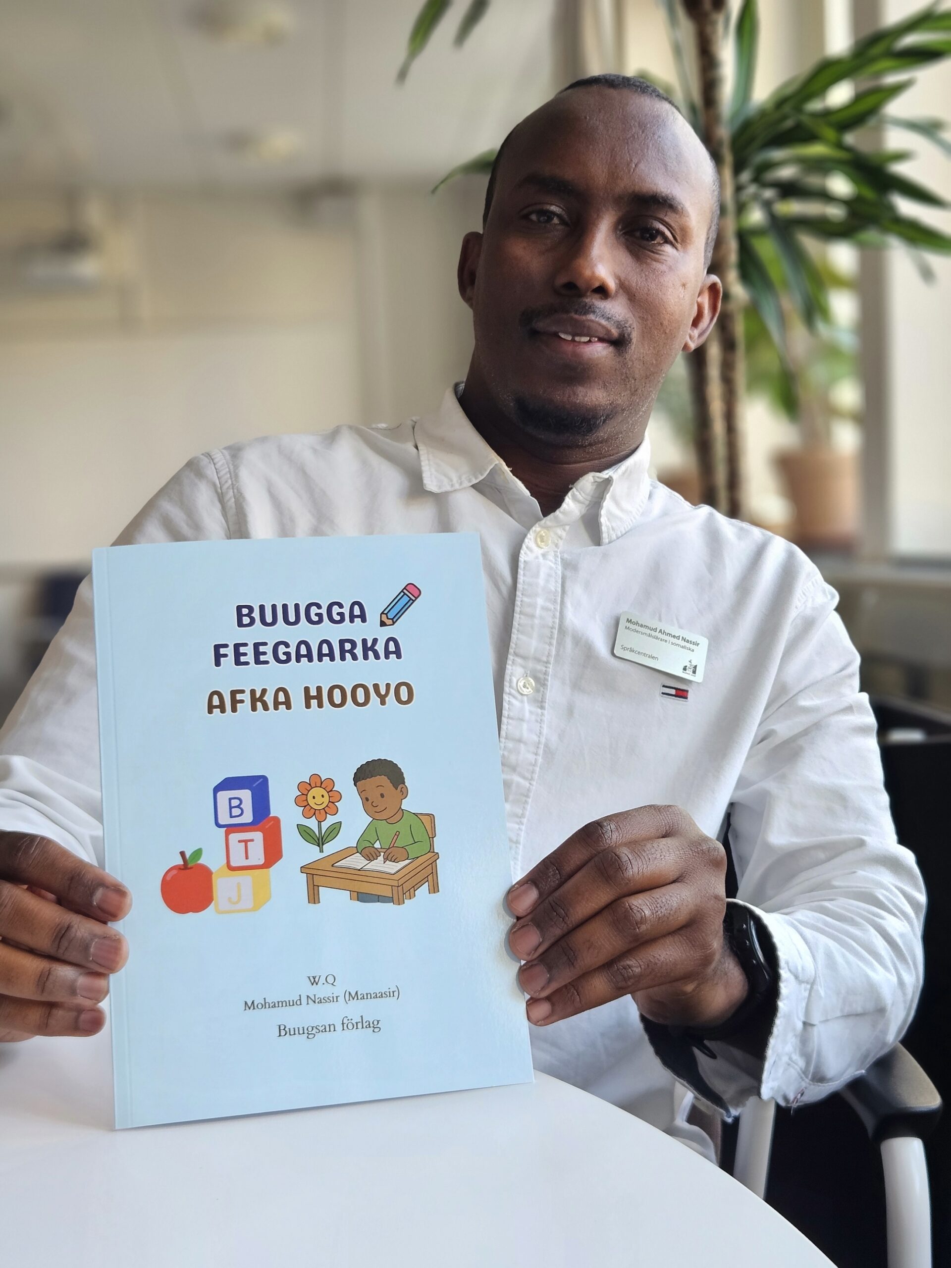 Person håller upp en ljusblå bok med titeln ”Buugga Feegaar ka Afka Hooyo” vid ett bord i ett ljust rum.