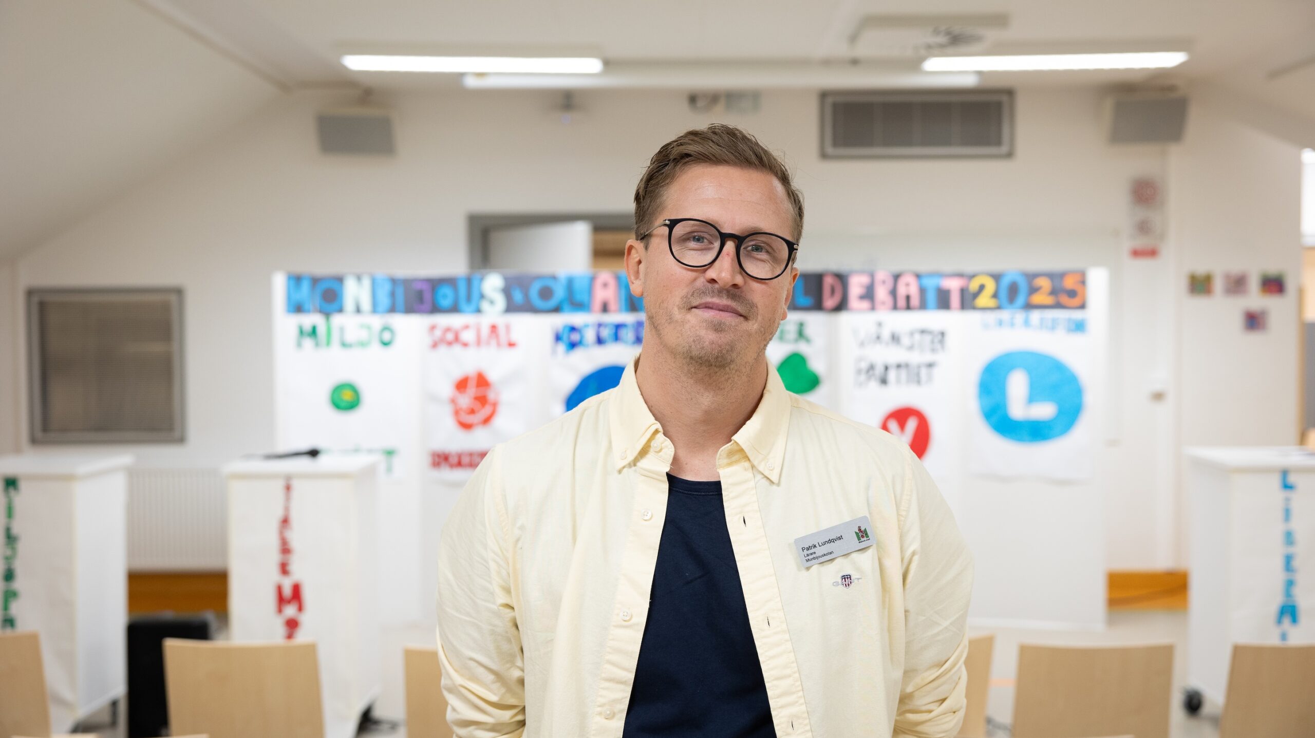 Person står i ett rum med stolar och affischer med politiska symboler och texten ”Miljö”, ”Social”, ”Identitet”, ”2025”.