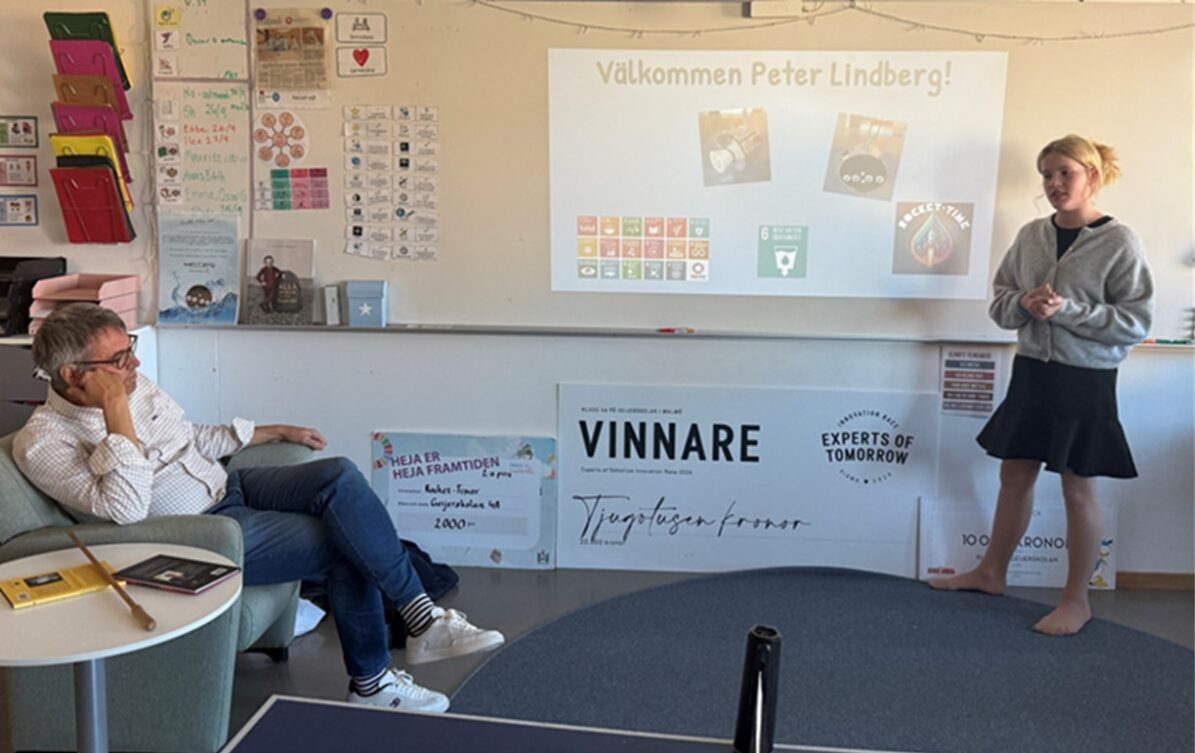 Person står vid en projektorbild med texten ”Välkommen Peter Lindberg” medan en annan sitter i en stol i klassrum.