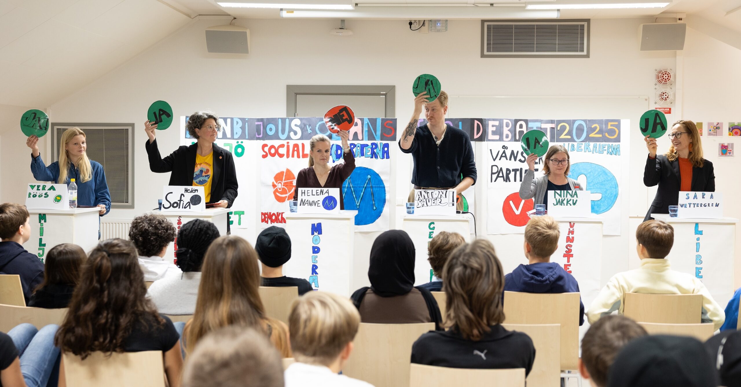 Skoldebatt med sex personer vid podium som håller upp gröna och röda skyltar inför en publik av elever.