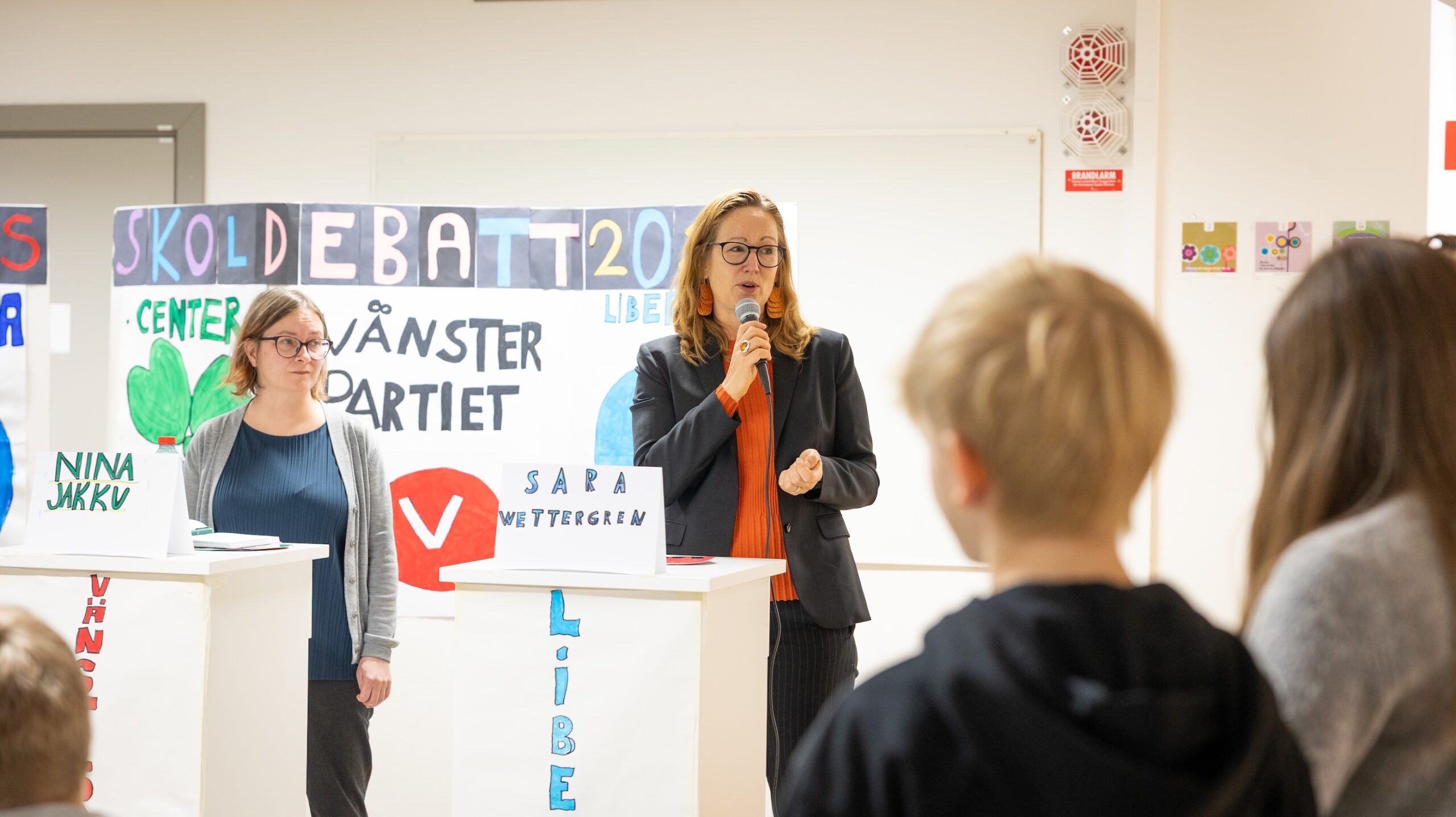 Två personer står vid talarpodium under en skoldebatt med affischer för olika partier, medan elever lyssnar.