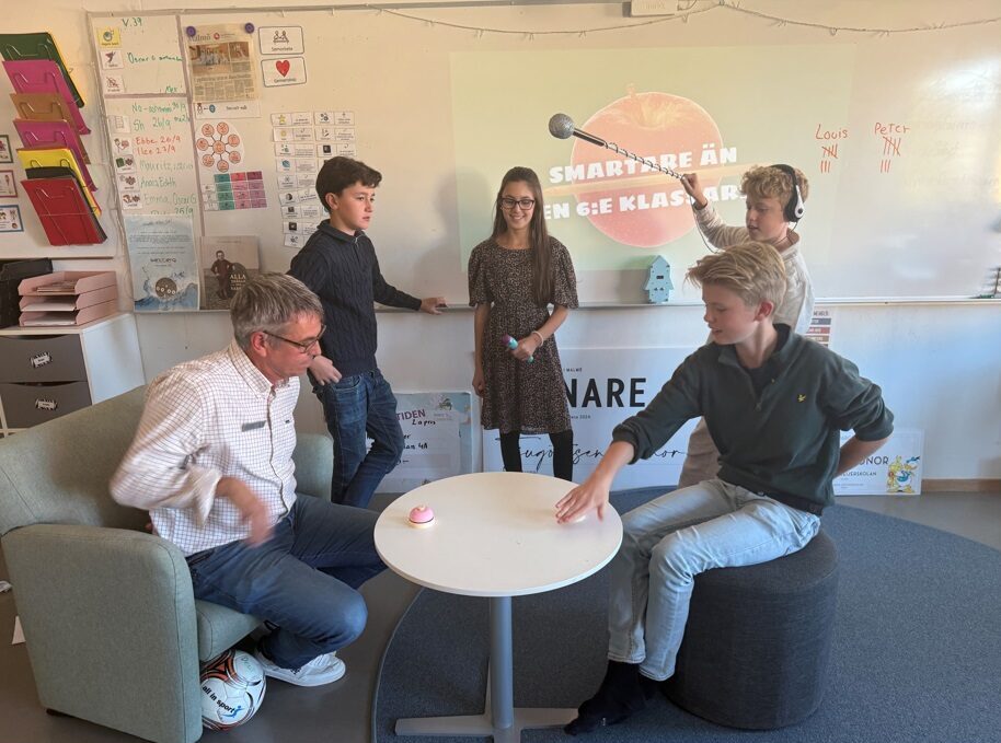 Grupp i klassrum deltar i aktivitet med mikrofon framför en projektorbild som visar texten ”Smartare än en 6:e klassare”.