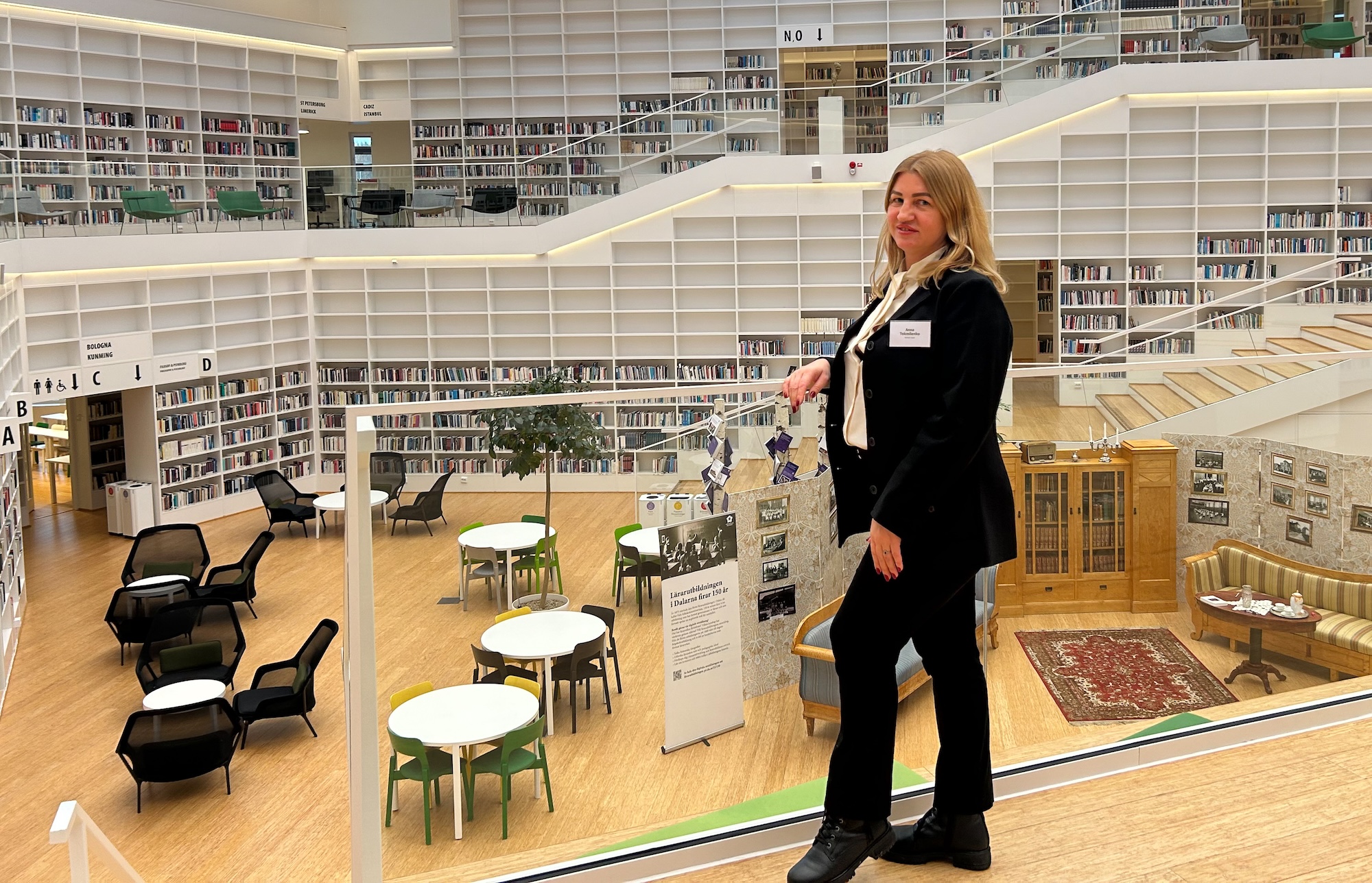 Modern biblioteksmiljö med flera våningar fyllda med böcker, öppna läsytor och sittgrupper. Bilden visar en person vid glasräcke med utsikt över bibliotekets ljusa interiör, inklusive bord, stolar och en utställning med historiska fotografier.