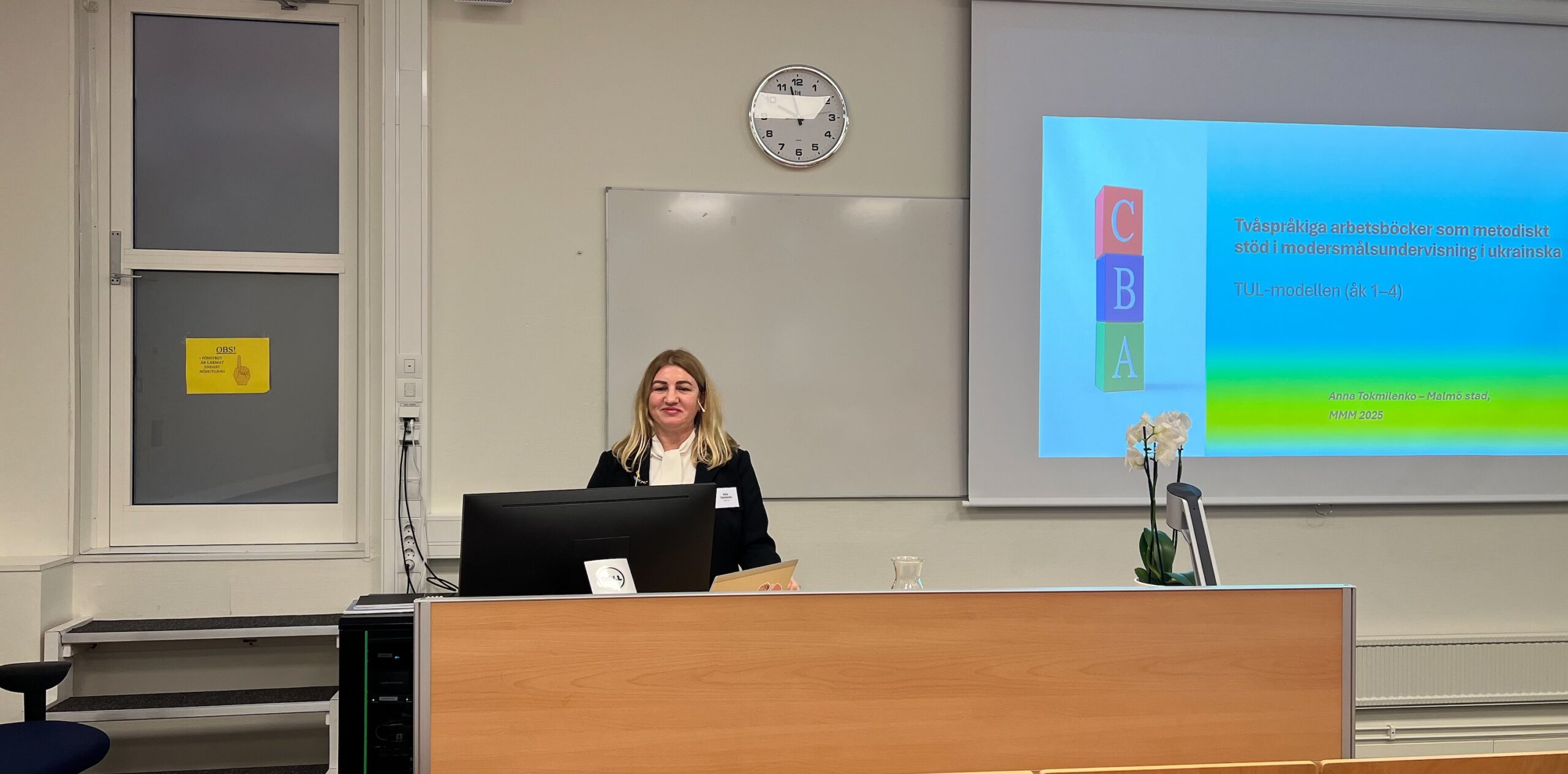 Föreläsning i klassrum med presentation om tvåspråkiga metoder inom matematikundervisning. Talare står vid en dator och projektorduk visar rubriken samt färgade block med bokstäverna C, B och A. En klocka syns på väggen och en vit orkidé står på talarbordet.