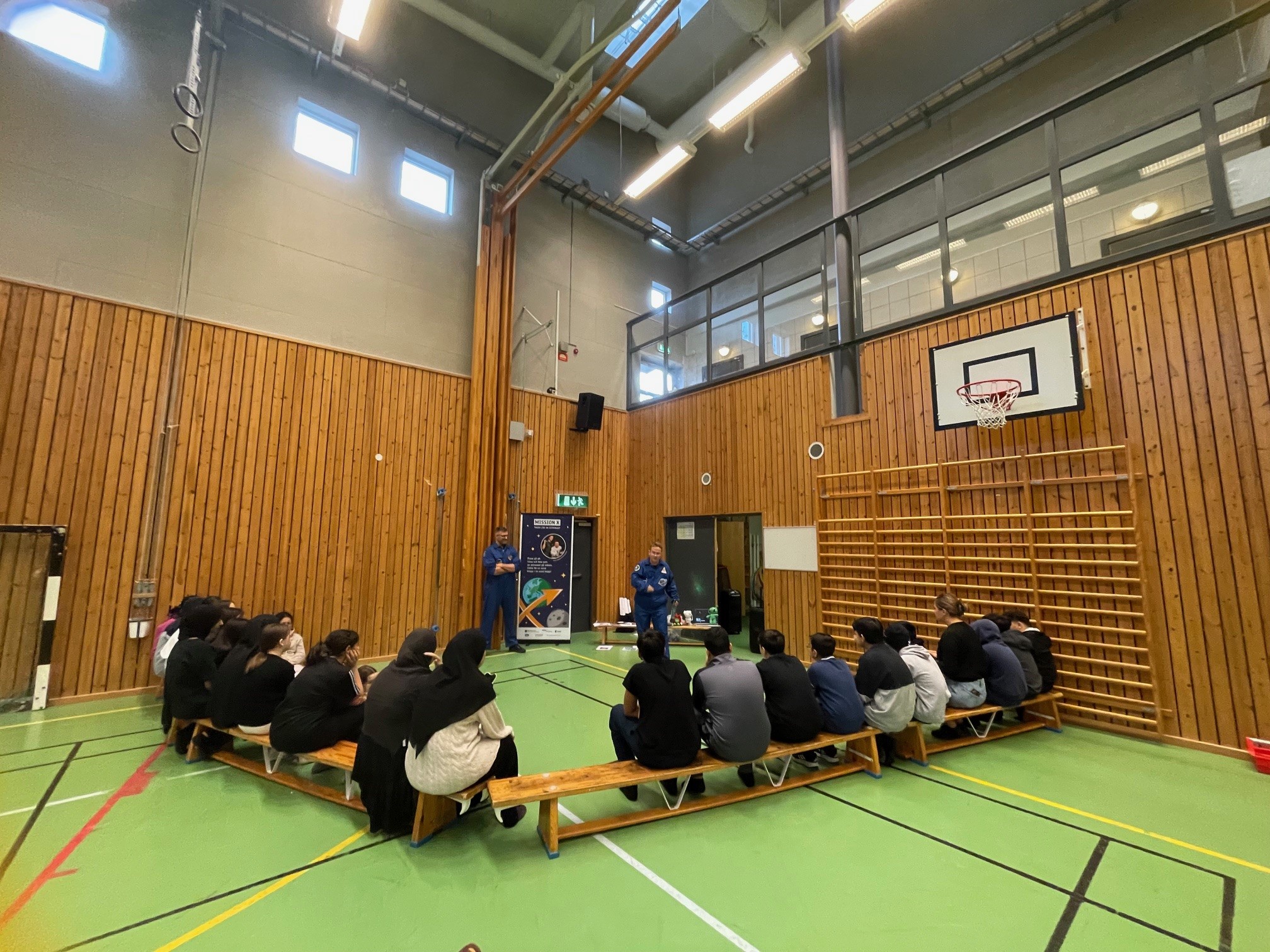 En grupp elever sitter på bänkar i en gymnastiksal med grönt golv och träpanelväggar, lyssnar på två personer i blå astronautdräkter som håller en rymdpresentation. En basketkorg och en affisch med rymdtema syns i bakgrunden.
