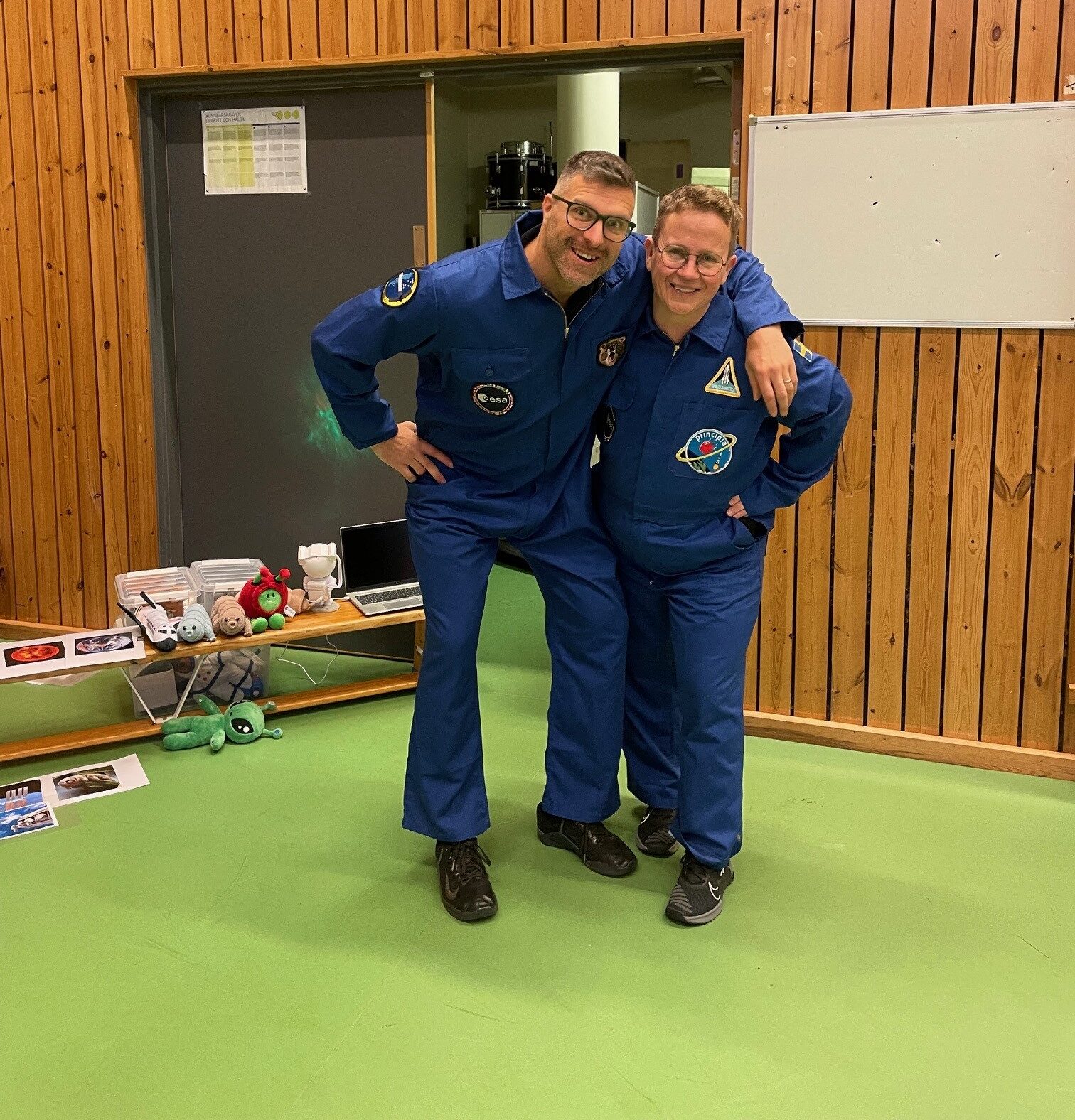 Två personer i blå astronautdräkter står i ett rum med träpanelväggar och grönt golv. Bakom dem syns en grå dörr, en vit anslagstavla och ett bord med gosedjur och rymdrelaterade föremål.