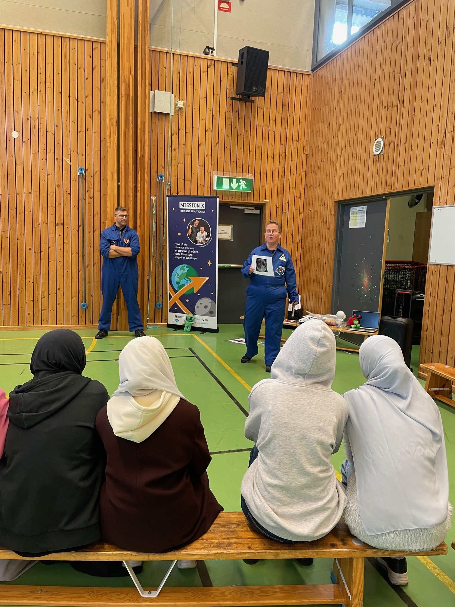 Fyra personer sitter på en bänk och lyssnar på två personer i blå astronautdräkter som håller en presentation i en gymnastiksal med träpanelväggar. En affisch med rymdtema står bredvid en dörr, och en av presentatörerna visar en bild.