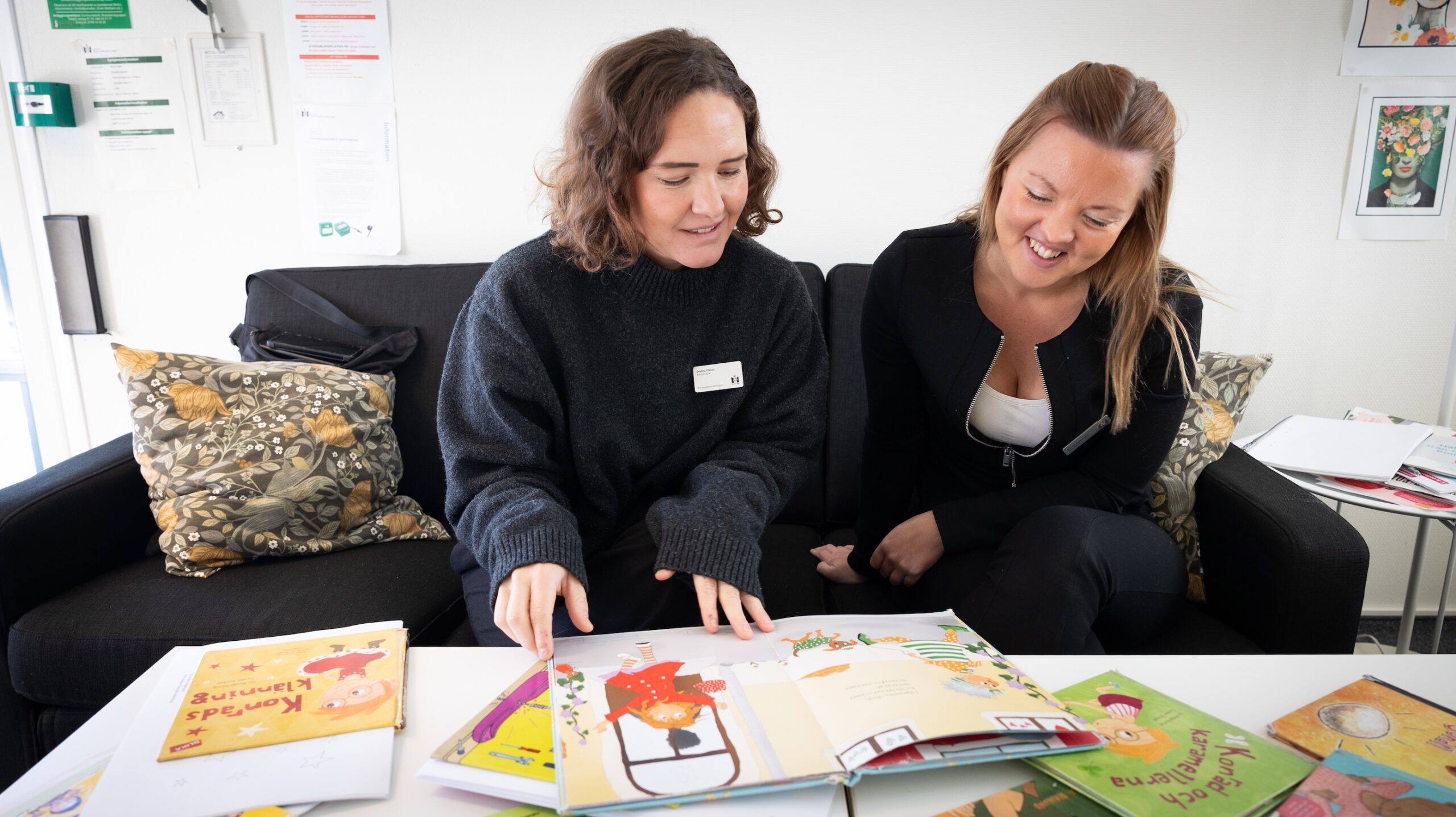Två personer sitter i en soffa och bläddrar i färgglada barnböcker på ett vitt bord. Runt dem ligger flera illustrerade böcker utspridda, och bakom syns dekorativa kuddar samt en ljus vägg med affischer.