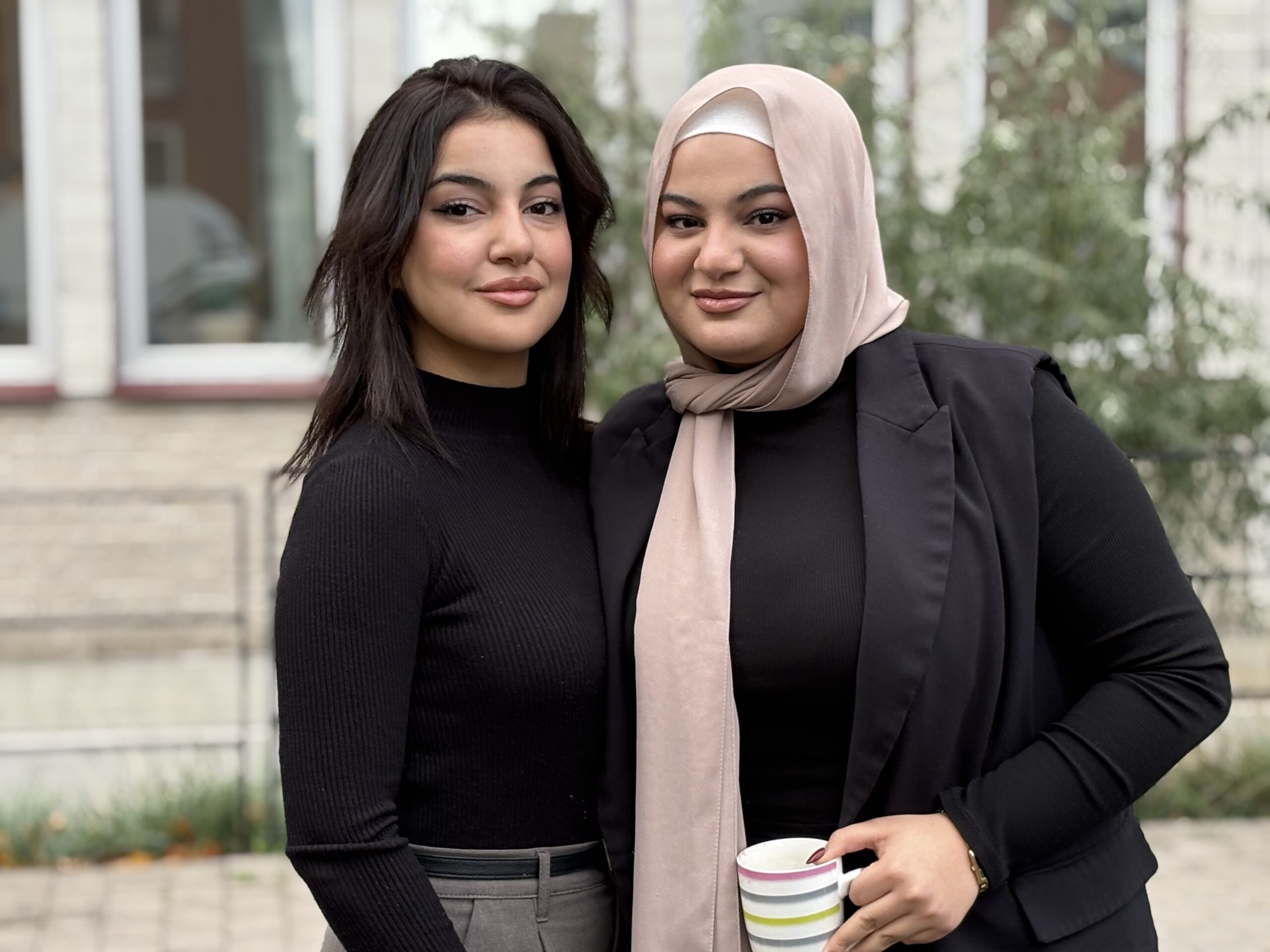 Två personer står nära varandra utomhus, en med beige sjal och randig mugg, den andra i svart tröja.