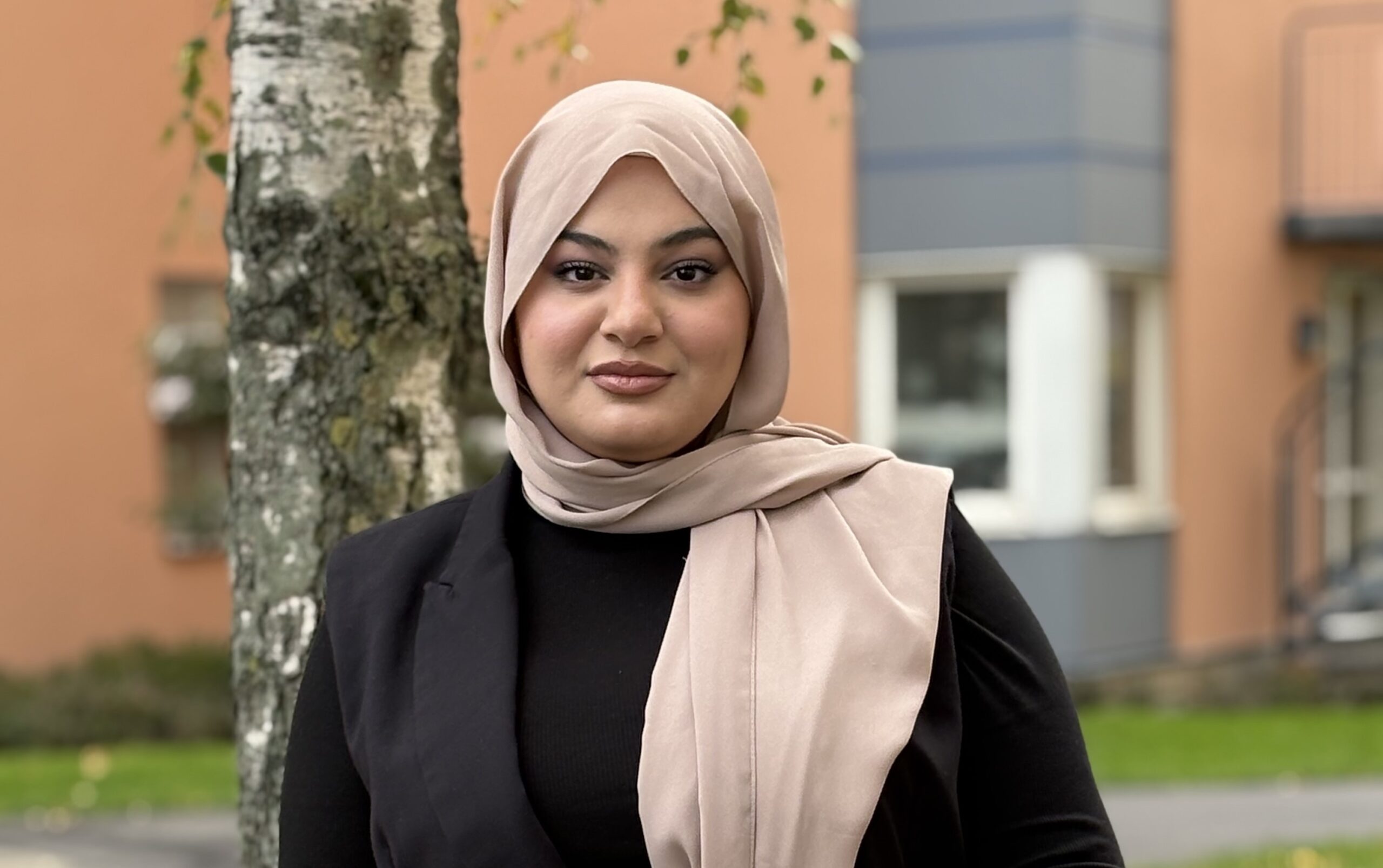 Person utomhus med beige sjal och svart kavaj, står bredvid ett träd med byggnad i bakgrunden.