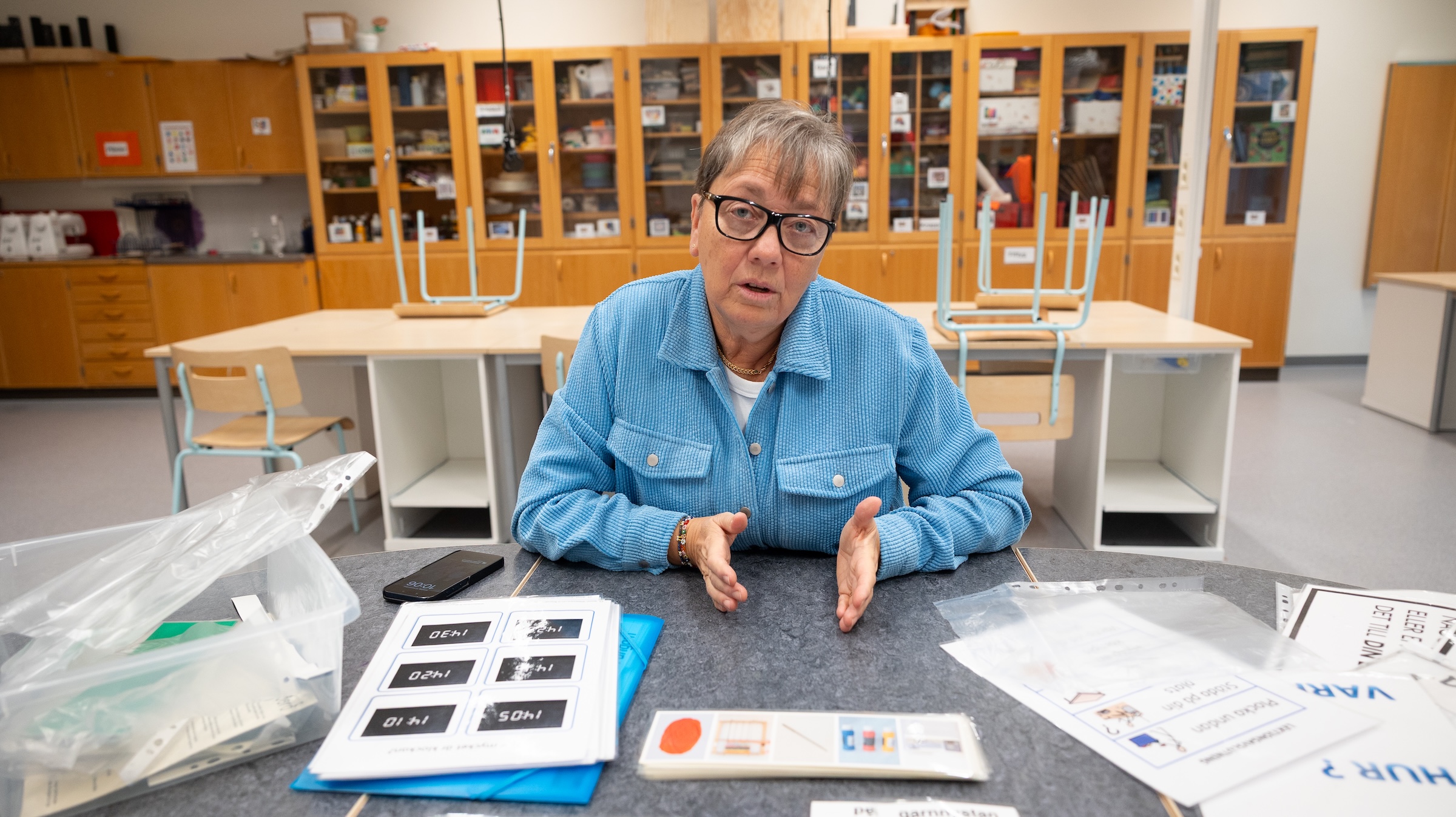 Monika Nilsson sitter vid ett bord i ett klassrum med träskåp och hyllor i bakgrunden. På bordet ligger plastfickor med instruktioner, bildkort och en mobiltelefon, vilket tyder på planering eller undervisning i textilslöjd.