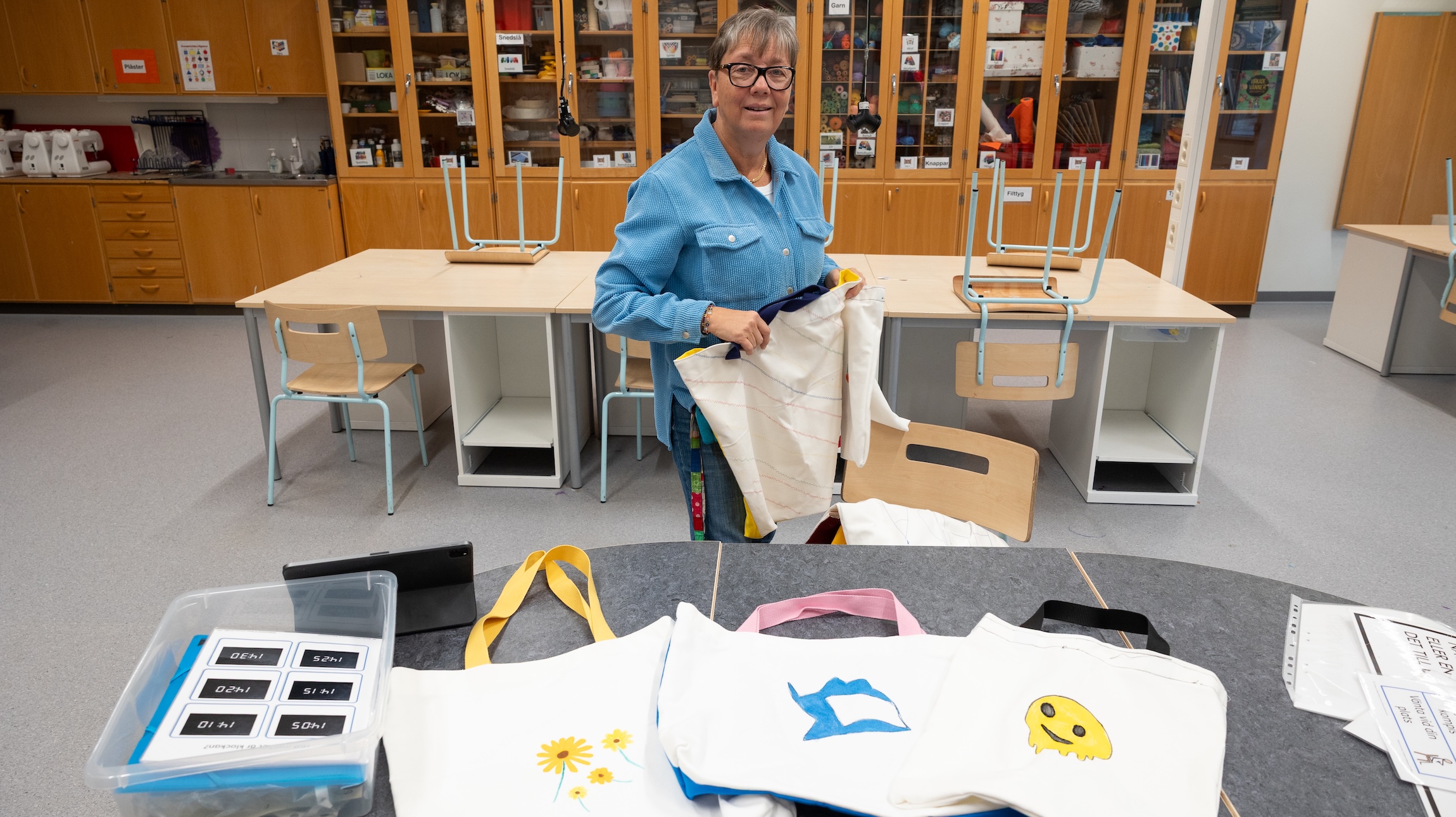 Monika Nilsson håller tygkassar i ett klassrum med träbord och hyllor fyllda med material. På bordet ligger flera handgjorda tygkassar med olika motiv, bland annat blommor, en blå form och en gul emoji.