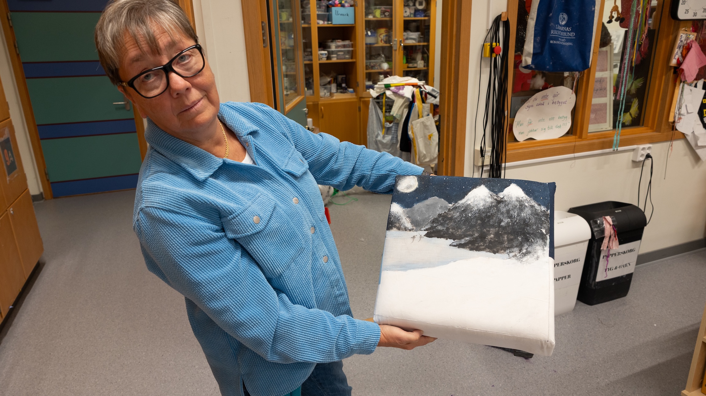 Monika Nilsson visar upp en målning med snötäckta berg och månsken, skapad på duk i ett klassrum med hyllor och hantverksmaterial i bakgrunden.