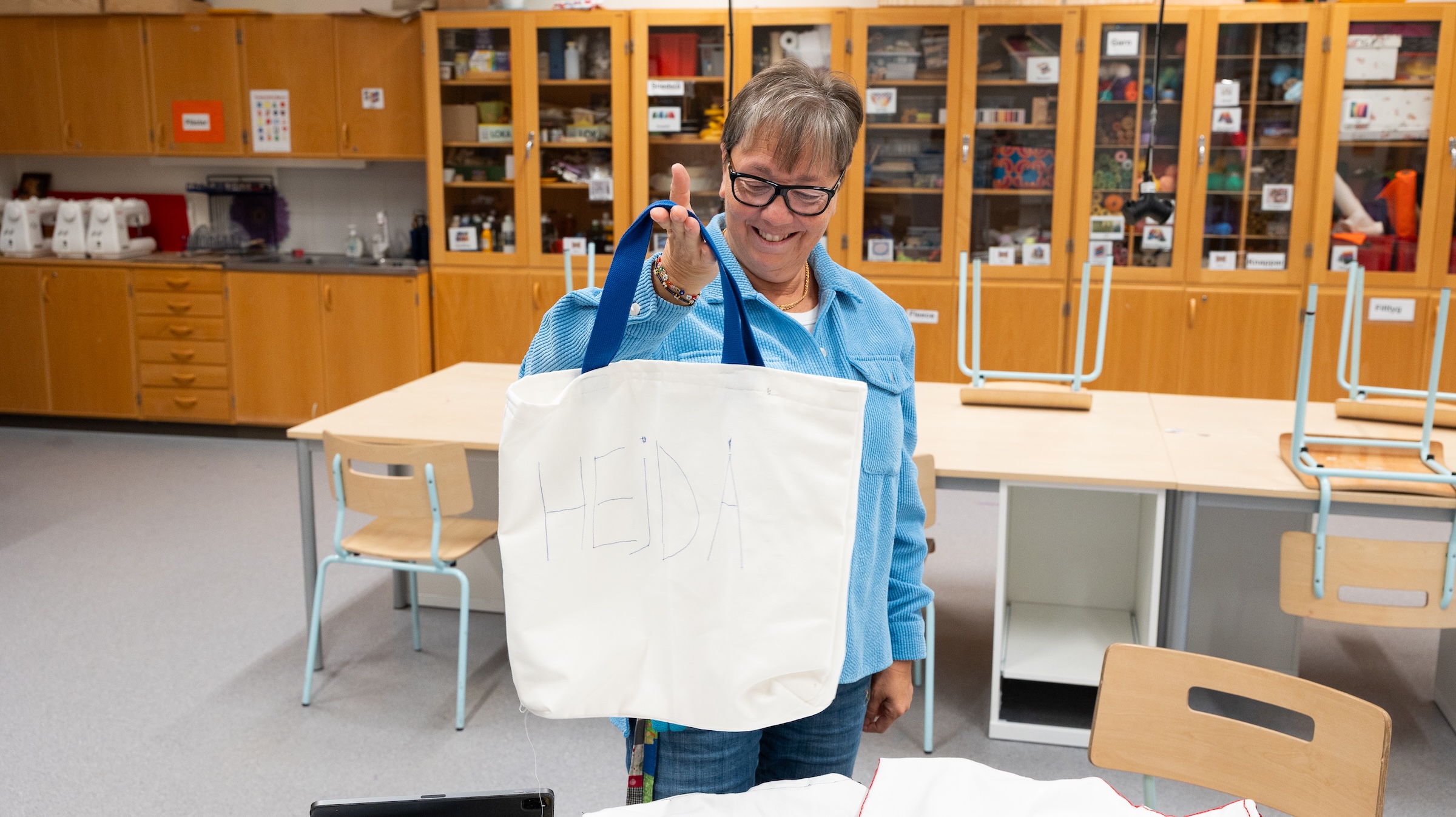 Monika Nilsson håller upp en vit tygkasse med blå handtag i ett klassrum med träbord och hyllor fyllda med material. På tygkassen står handskrivet 'hej då'.