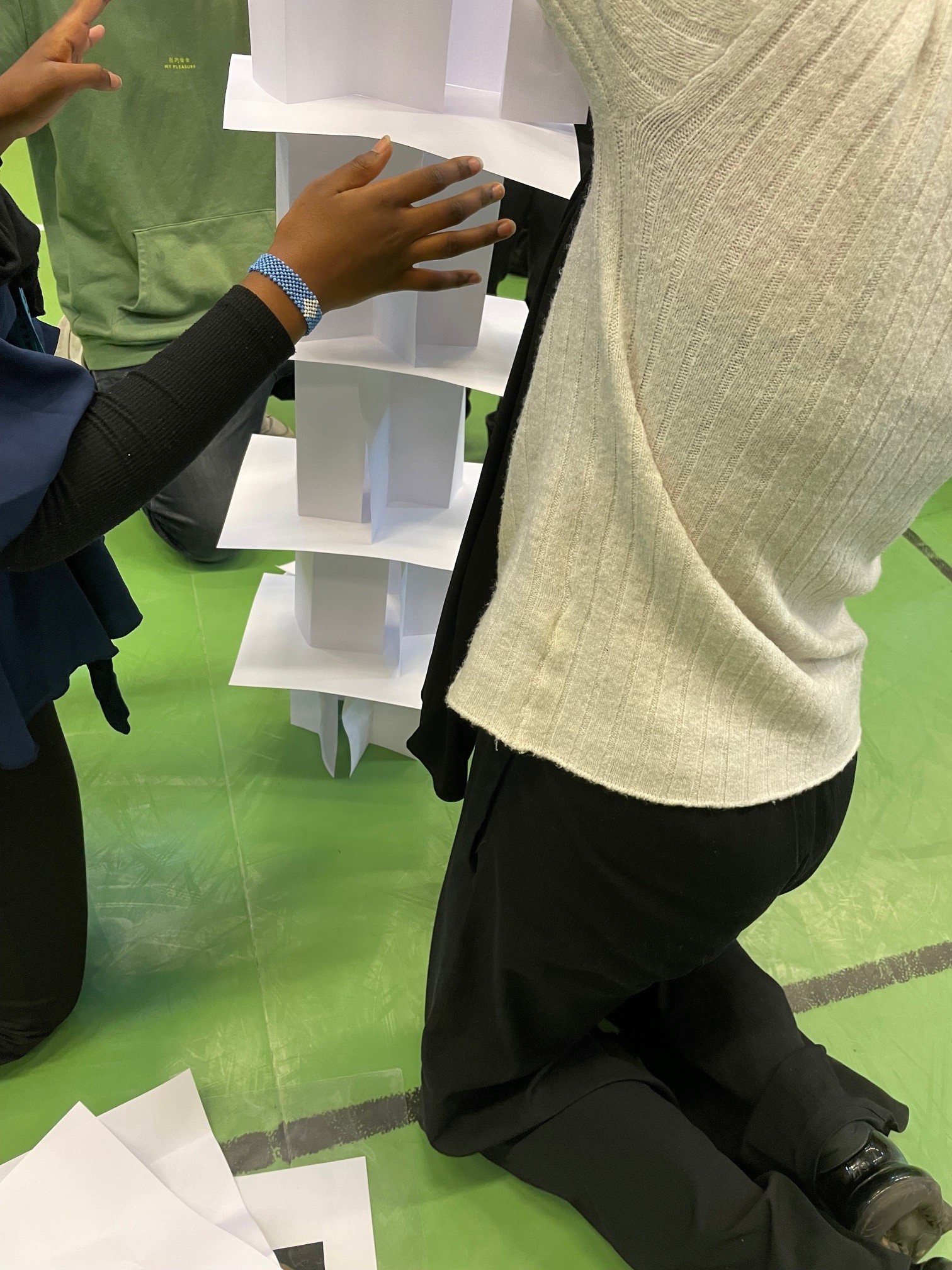 Två personer bygger en hög struktur av vita pappersark på ett grönt golv, som en del av en kreativ aktivitet eller workshop.