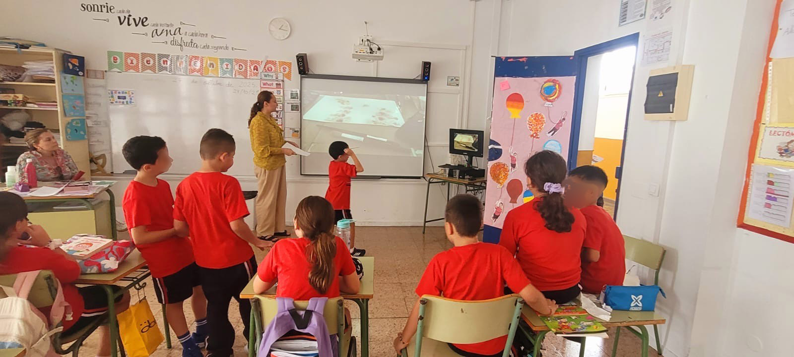 Barn i röda tröjor sitter och står i ett klassrum medan en person vid projektorduken visar något på skärmen. Klassrummet har färgglada dekorationer, affischer och skolmaterial på borden.