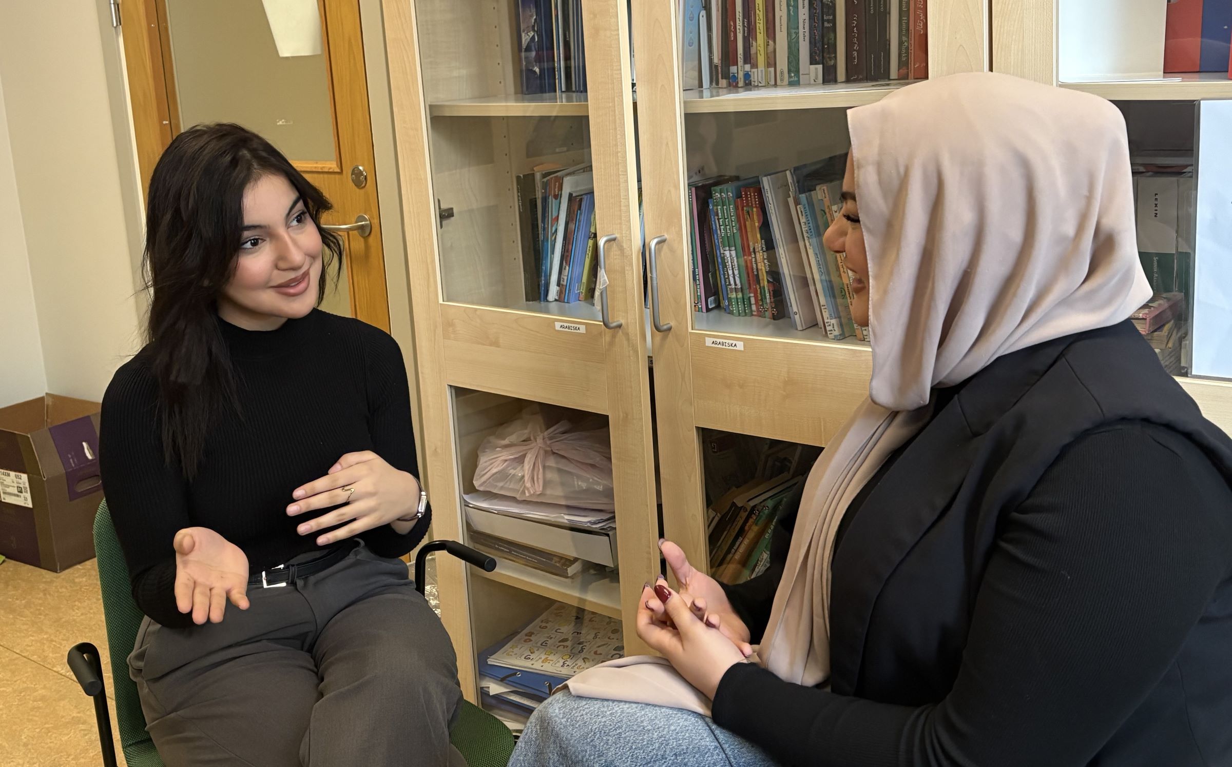 Två personer sitter inomhus och pratar, framför ett vitrinskåp med böcker.