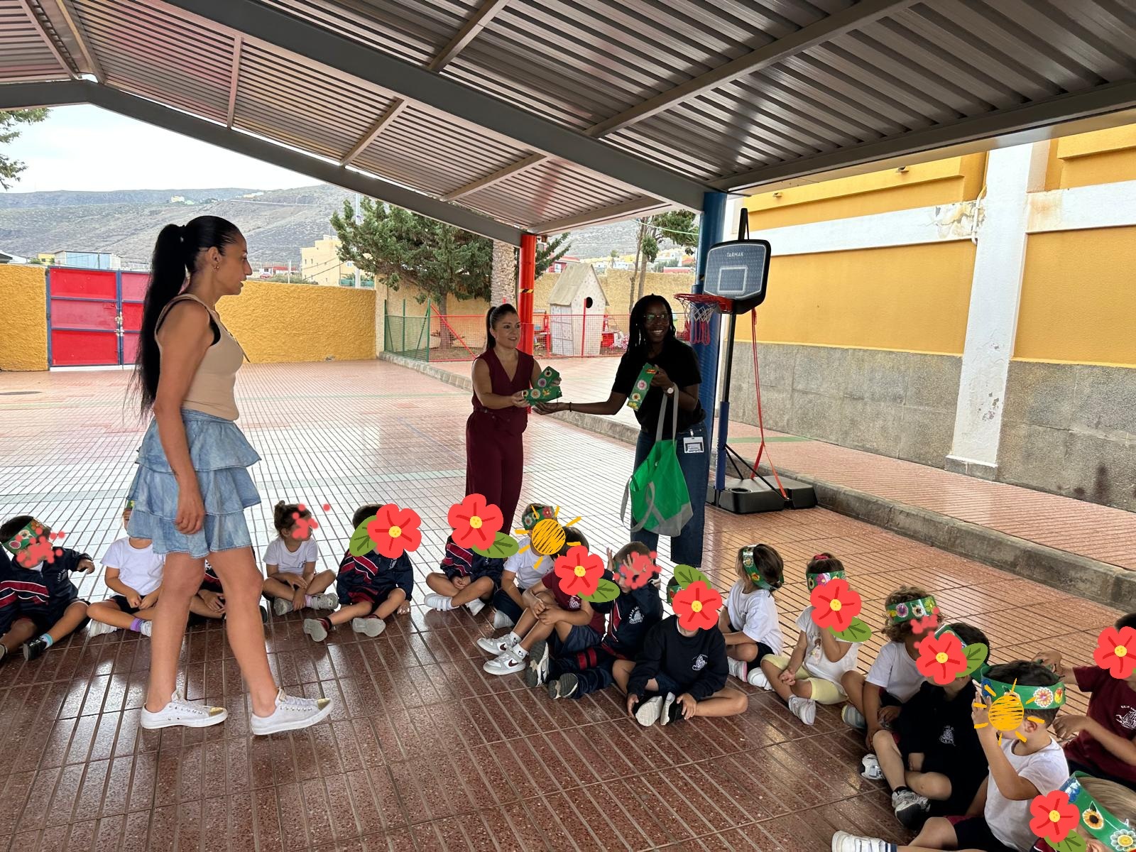 Två vuxna personer står under ett tak på en skolgård och håller i gröna föremål, medan en grupp barn sitter på marken framför dem i en halvcirkel. Barnen bär skoluniformer och har dekorerade papperskronor på huvudet.