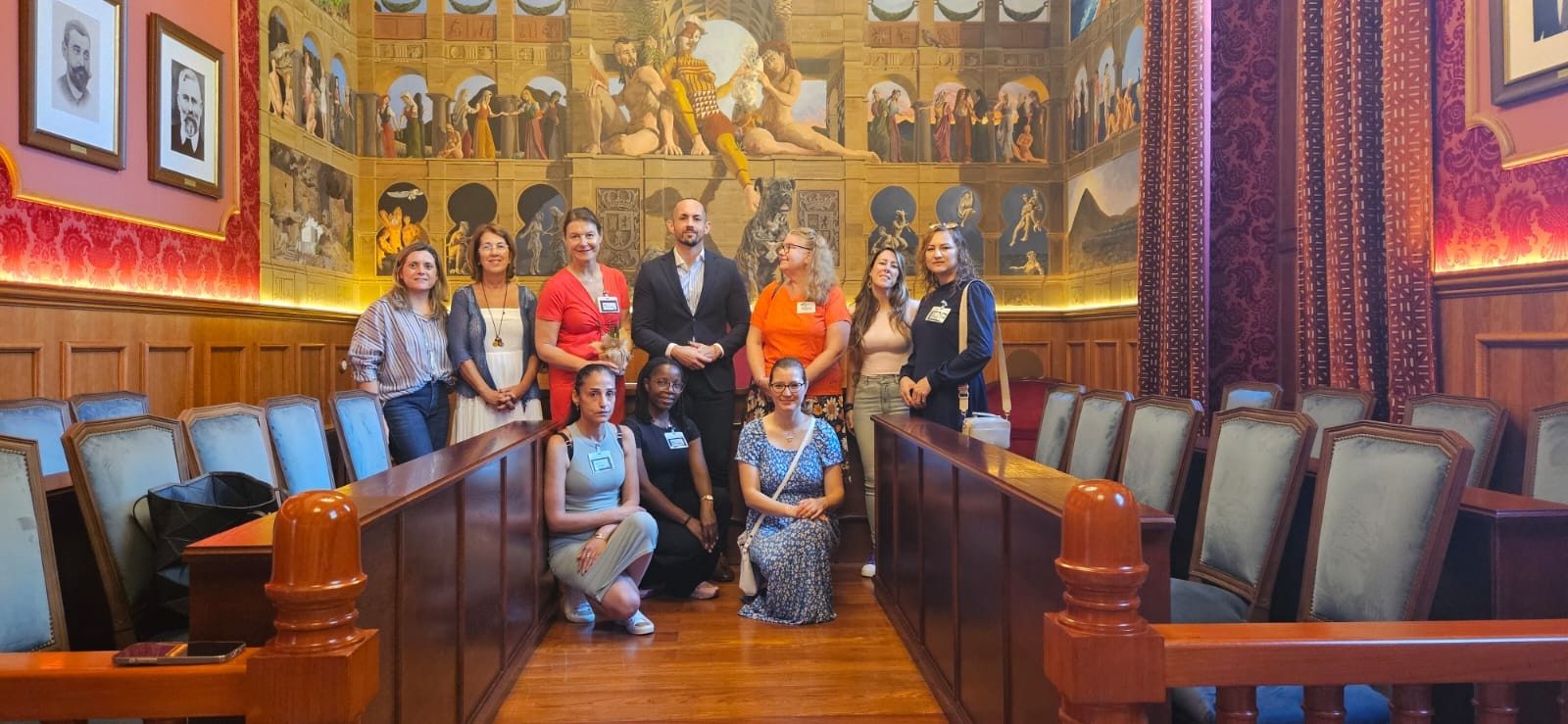 En grupp på nio personer poserar i ett historiskt mötesrum med träpaneler och röda väggar. Bakom gruppen syns en stor väggmålning med klassiska motiv och porträtt. Personerna står och sitter mellan två rader av mörka trästolar med gröna dynor.