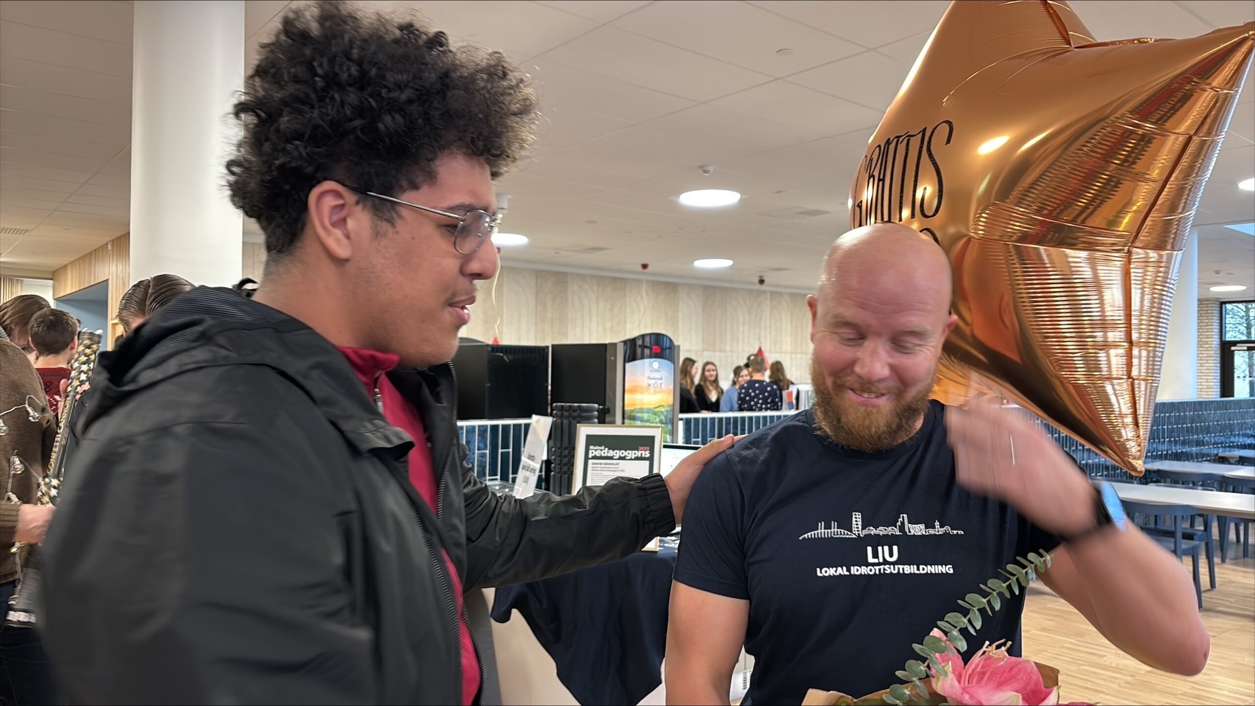 Två personer vid ett bord med guldfärgad stjärnballong och texten ”GRATTIS” under ett evenemang.