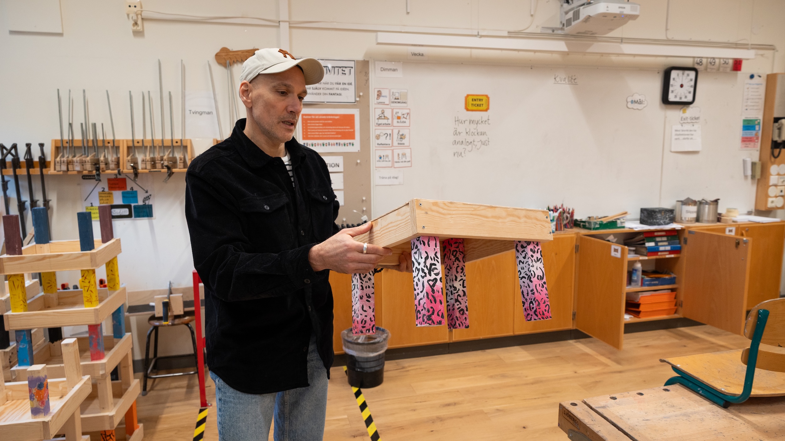 Person i slöjdsal håller i en fotpall med färgglada mönstrade band. Bakgrunden visar hyllor med verktyg och arbetsytor i skolmiljö för träslöjd.