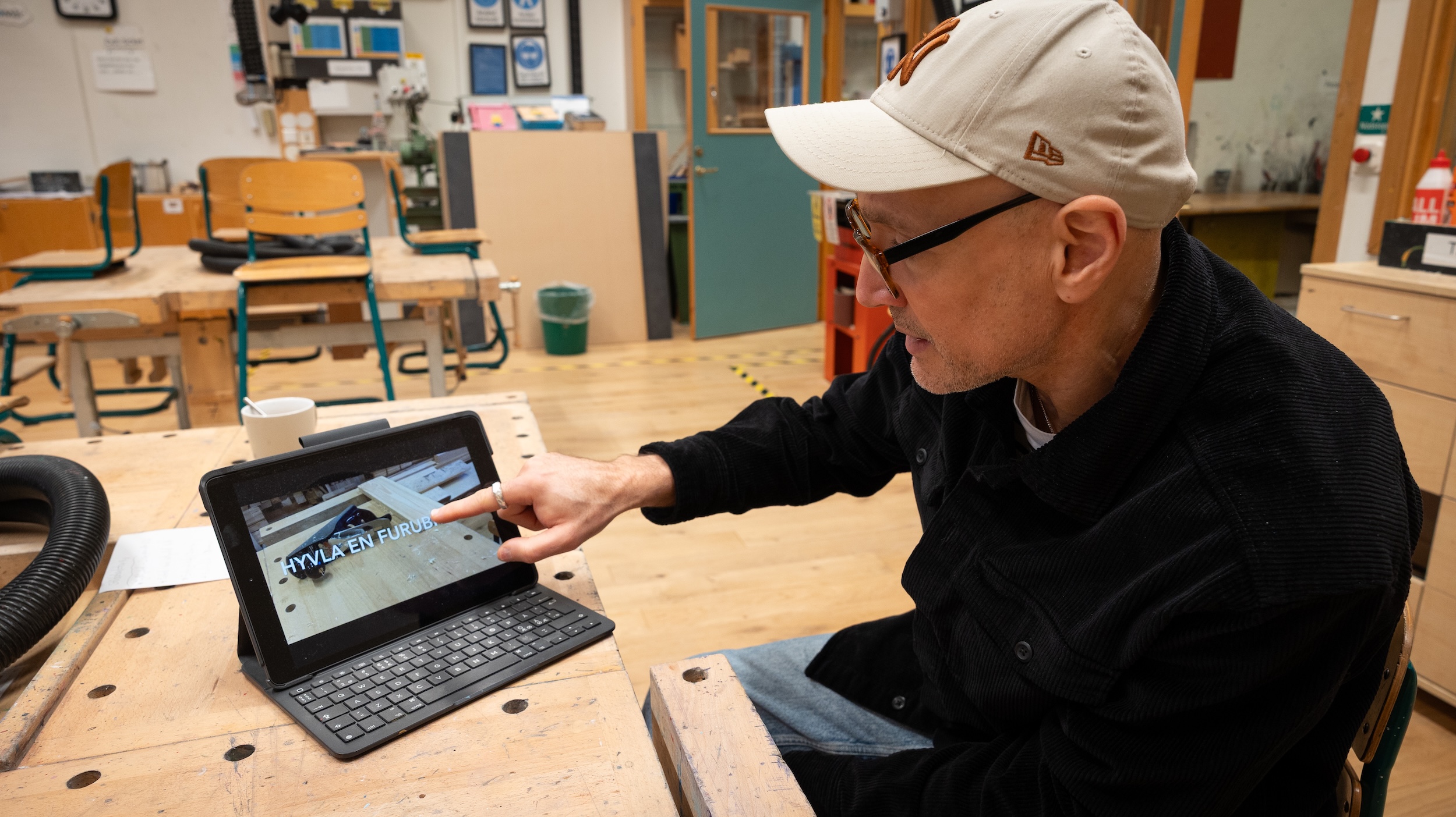Person i slöjdsal pekar på en surfplatta med bild av träprojekt. Bakgrunden visar arbetsbänkar, stolar och verktyg i skolmiljö för träslöjd.