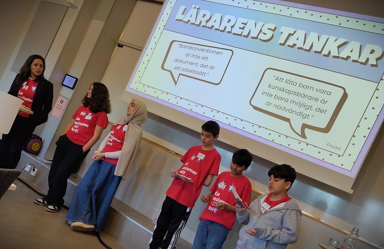Grupp av elever står framför en projektorduk under en presentation på konferensen om demokrati och mänskliga rättigheter. På skärmen visas citat om barnkonventionen och vikten av att låta barn vara kunskapsbärare.