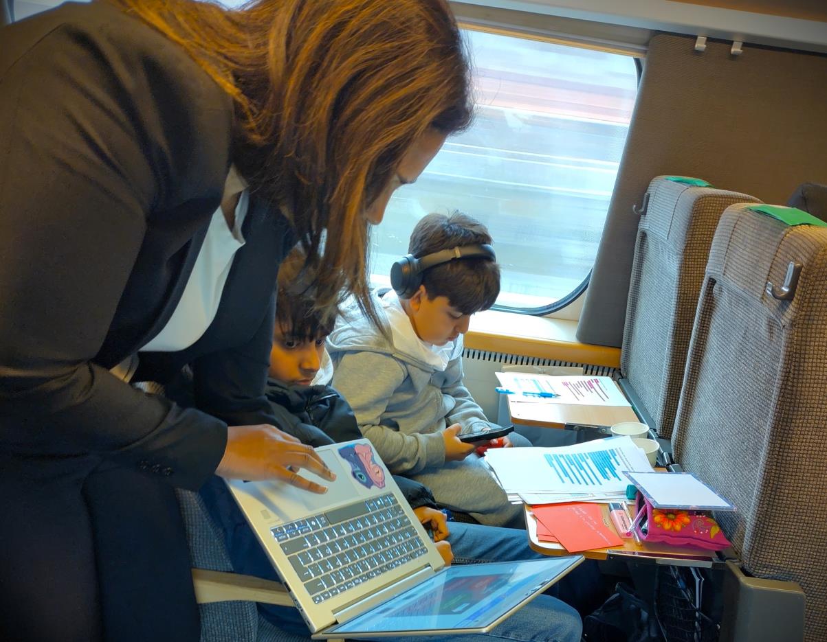 Person visar information på en bärbar dator för två elever som sitter på ett tåg med anteckningar och material på bordet, under resa till konferens om demokrati och mänskliga rättigheter.