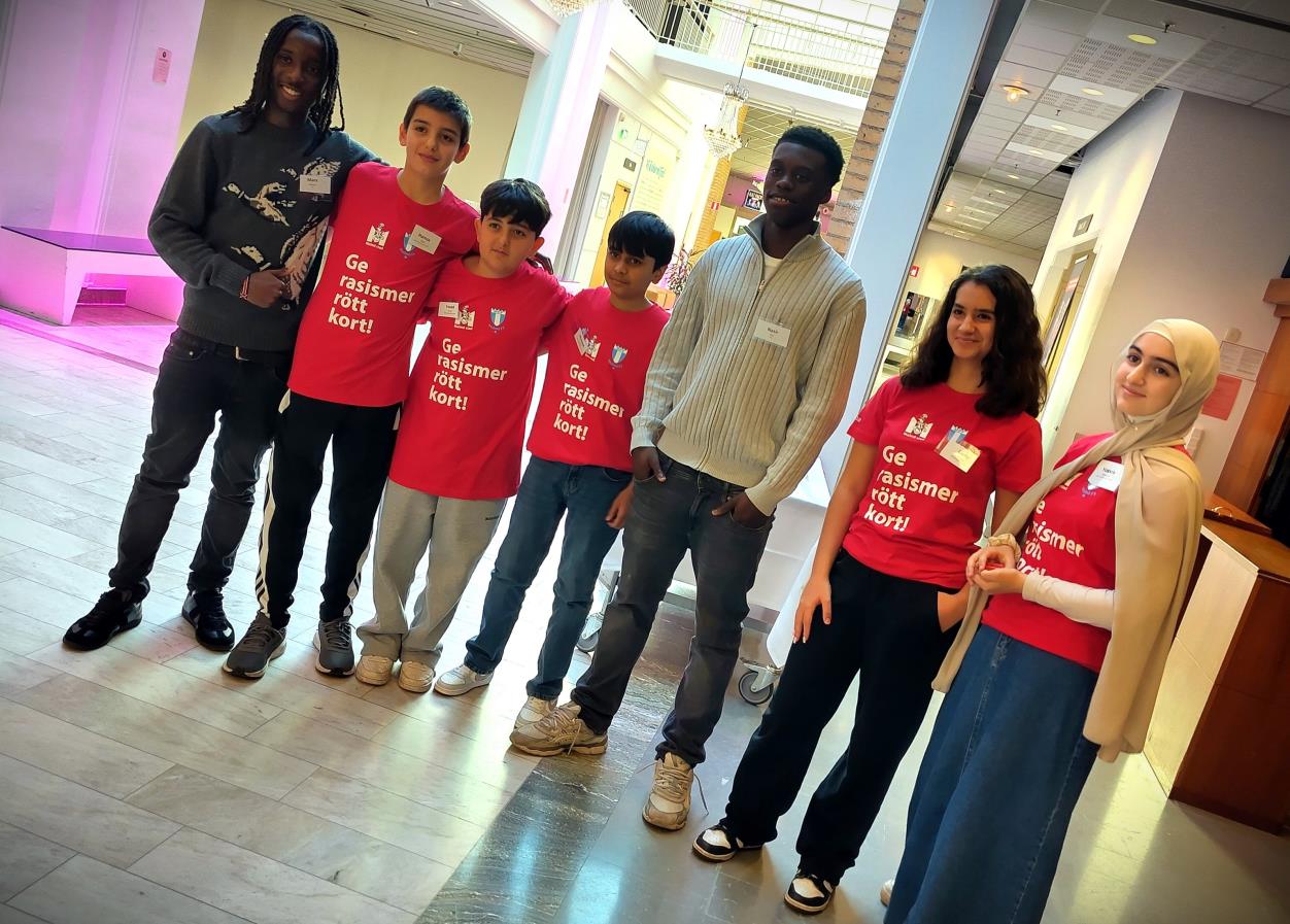 Grupp av elever och vuxna står tillsammans i en ljus konferensmiljö, flera bär röda tröjor med text om demokrati och mänskliga rättigheter.