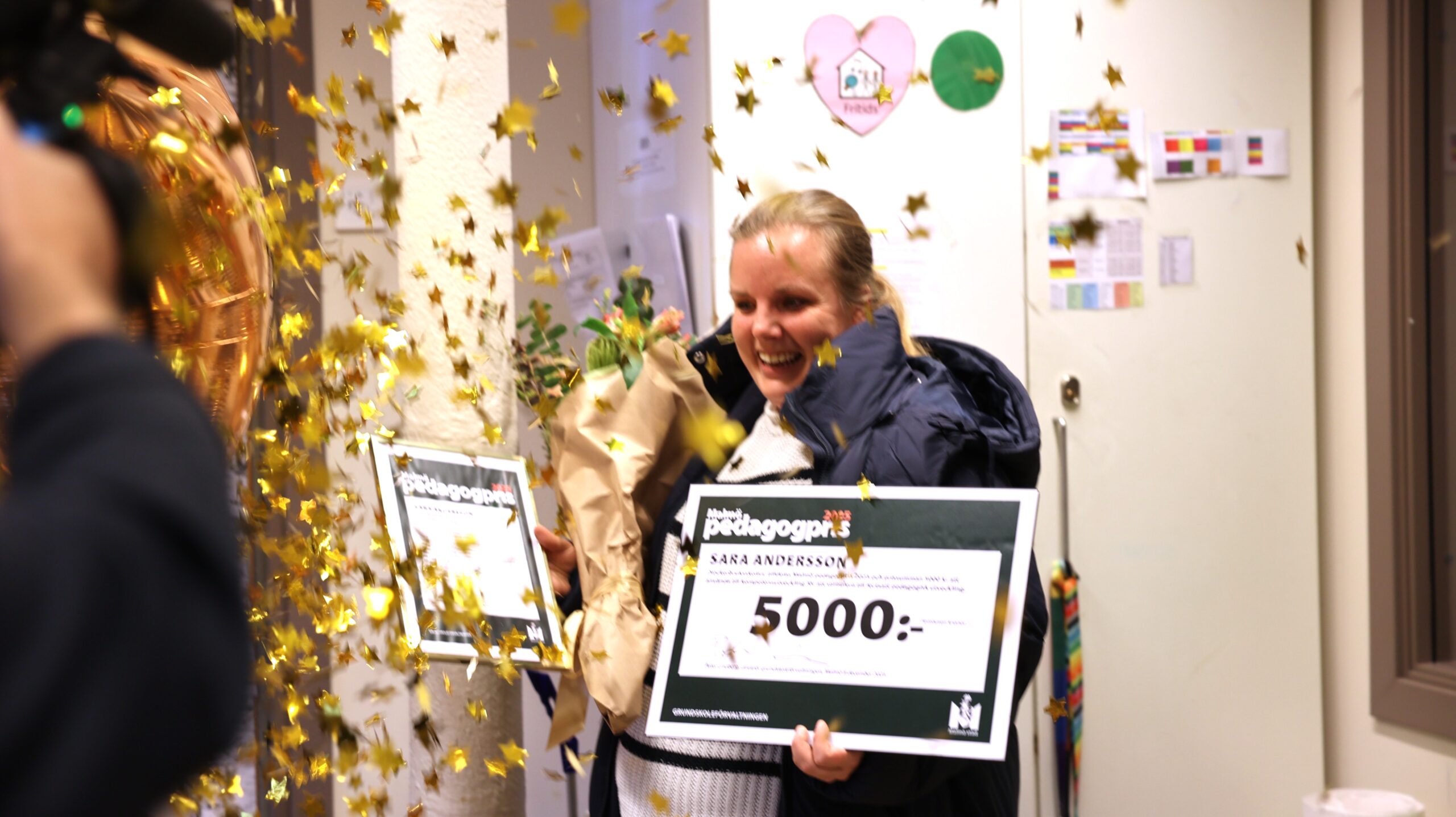 Pedagogprisvinnare står med check på 5000 kr och blombukett i konfettiregn.
