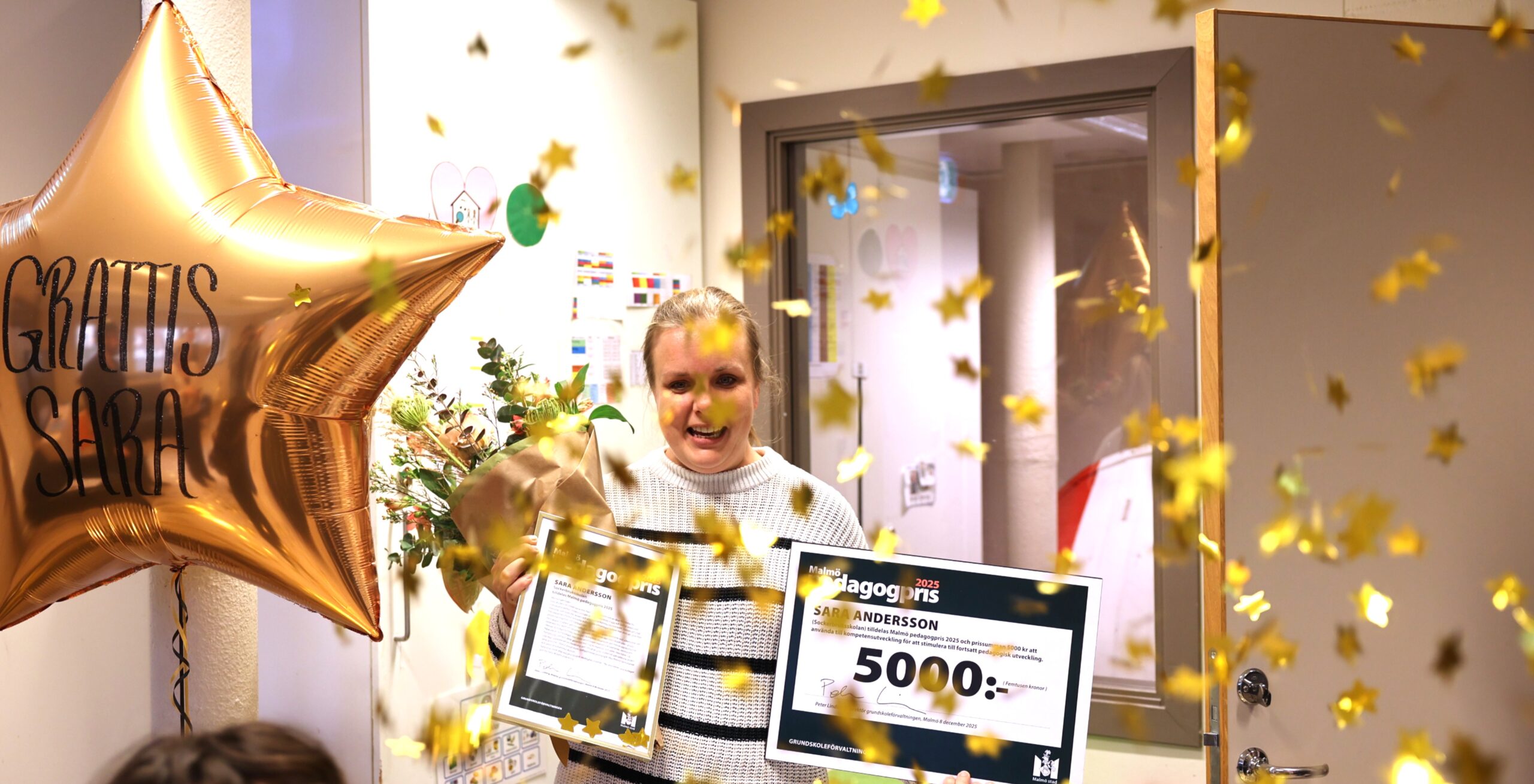Firande av Pedagogpriset med konfetti, blommor och en stor guldfärgad ballong med texten 'Grattis Sara'. Ett diplom och presentkort på 5000 kronor från Malmö stad syns tydligt i bilden.'