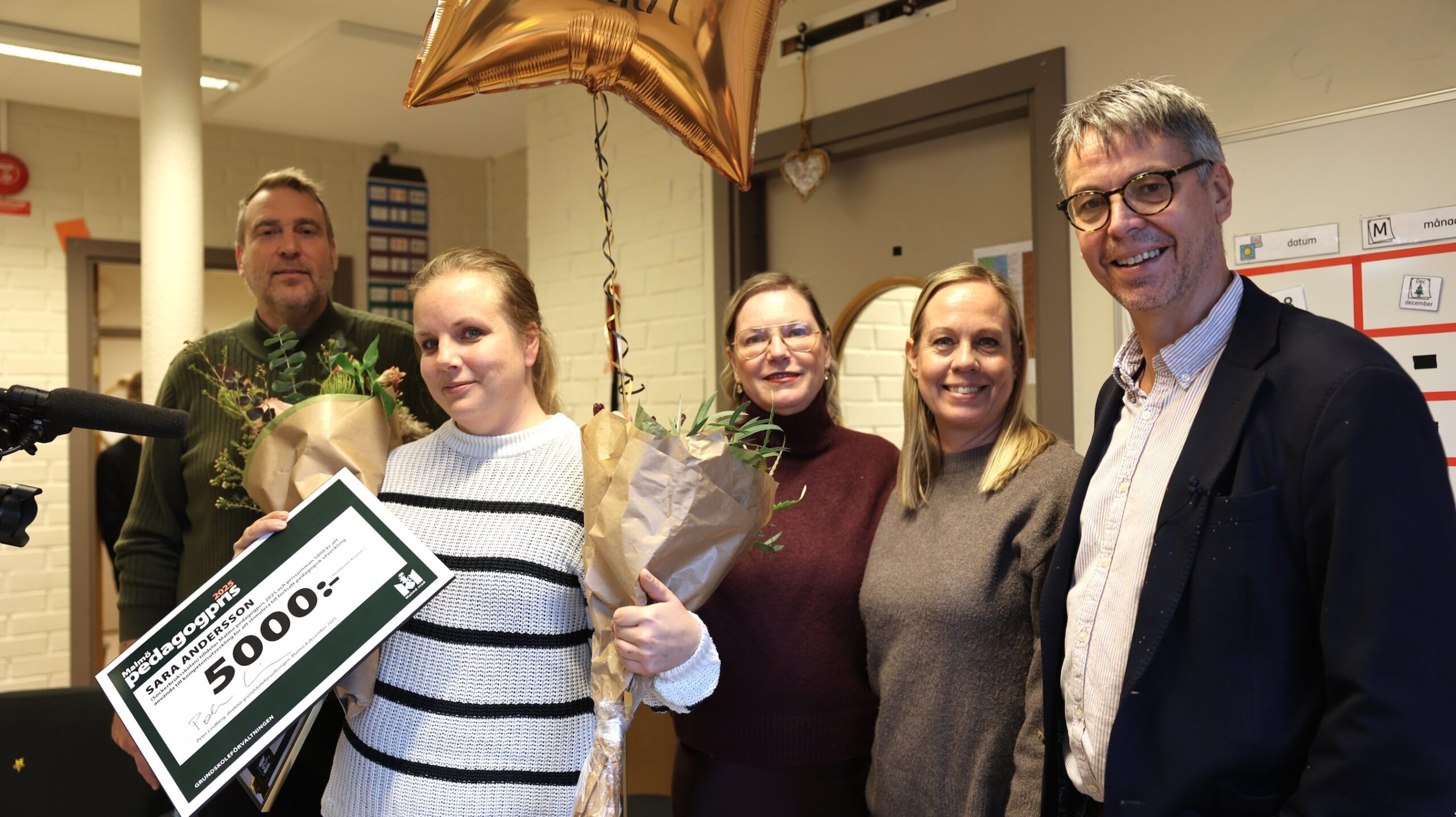 Grupp med personer i ett klassrumsmiljö firar med blommor, ballong och ett stort presentkort på 5000 kronor från Malmö stad.