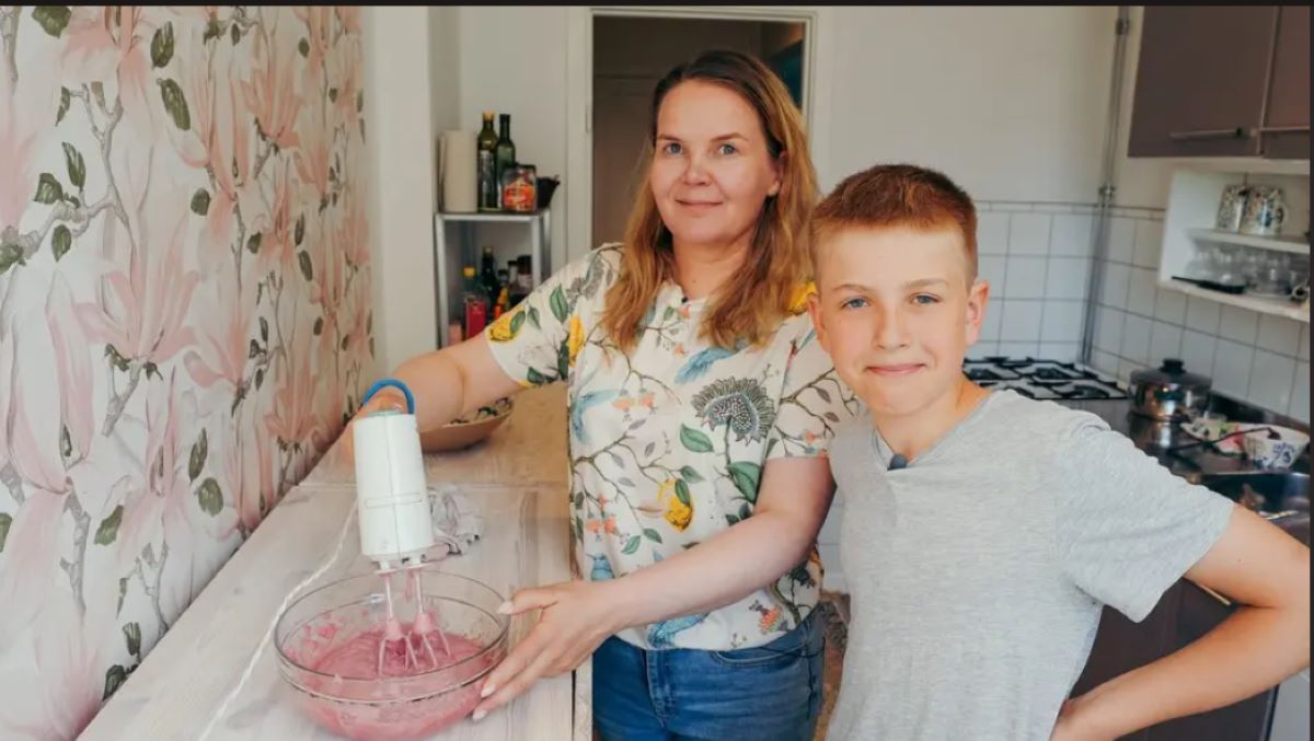 Person använder elvisp för att blanda rosa smet i en skål på köksbänk, med ett barn bredvid i ett ljust kök med blommig tapet.