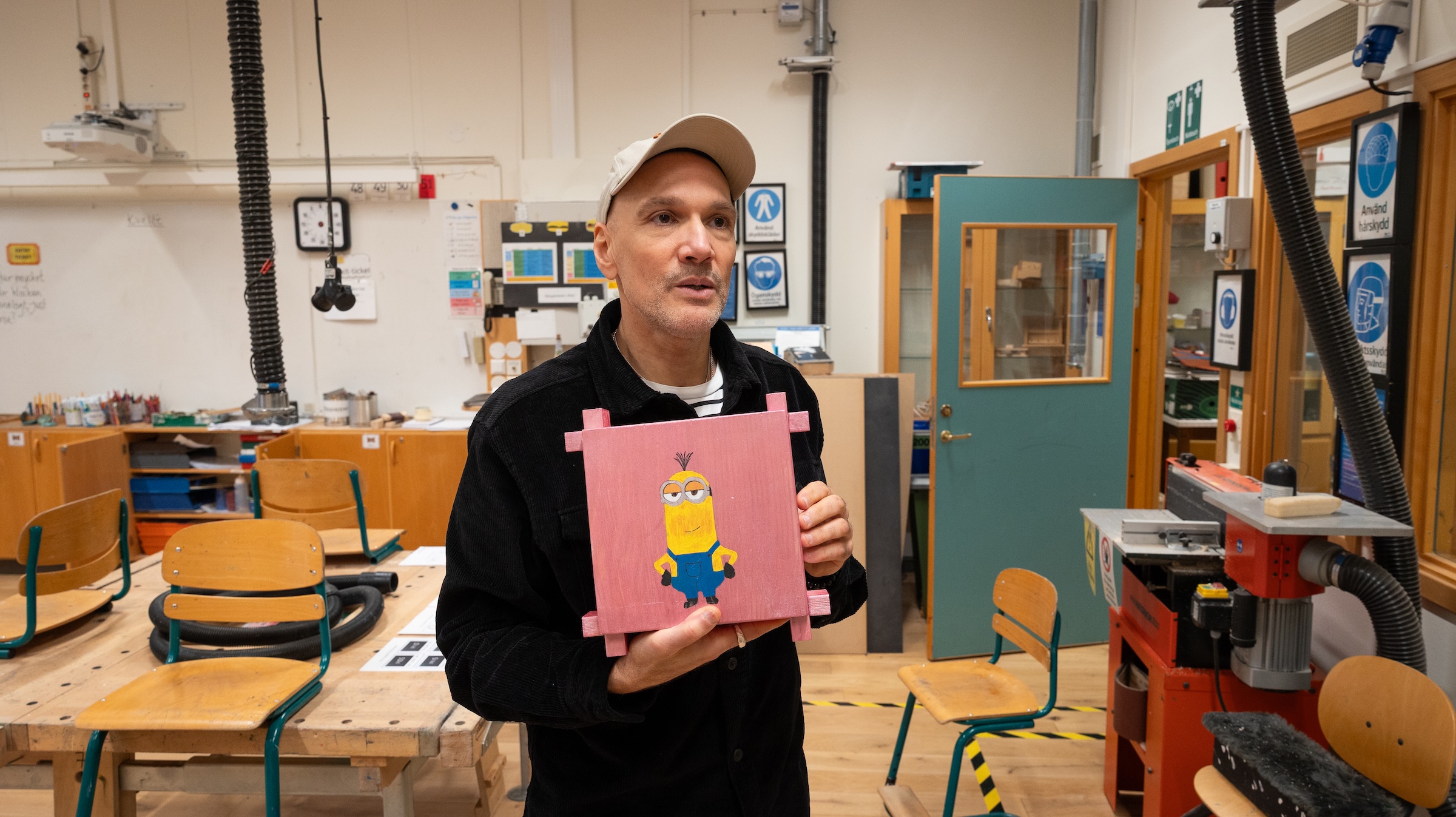Person i slöjdsal håller i en träskylt med målad Minion-figur. Bakgrunden visar arbetsbänkar, stolar och verktyg i en skolmiljö för träslöjd.