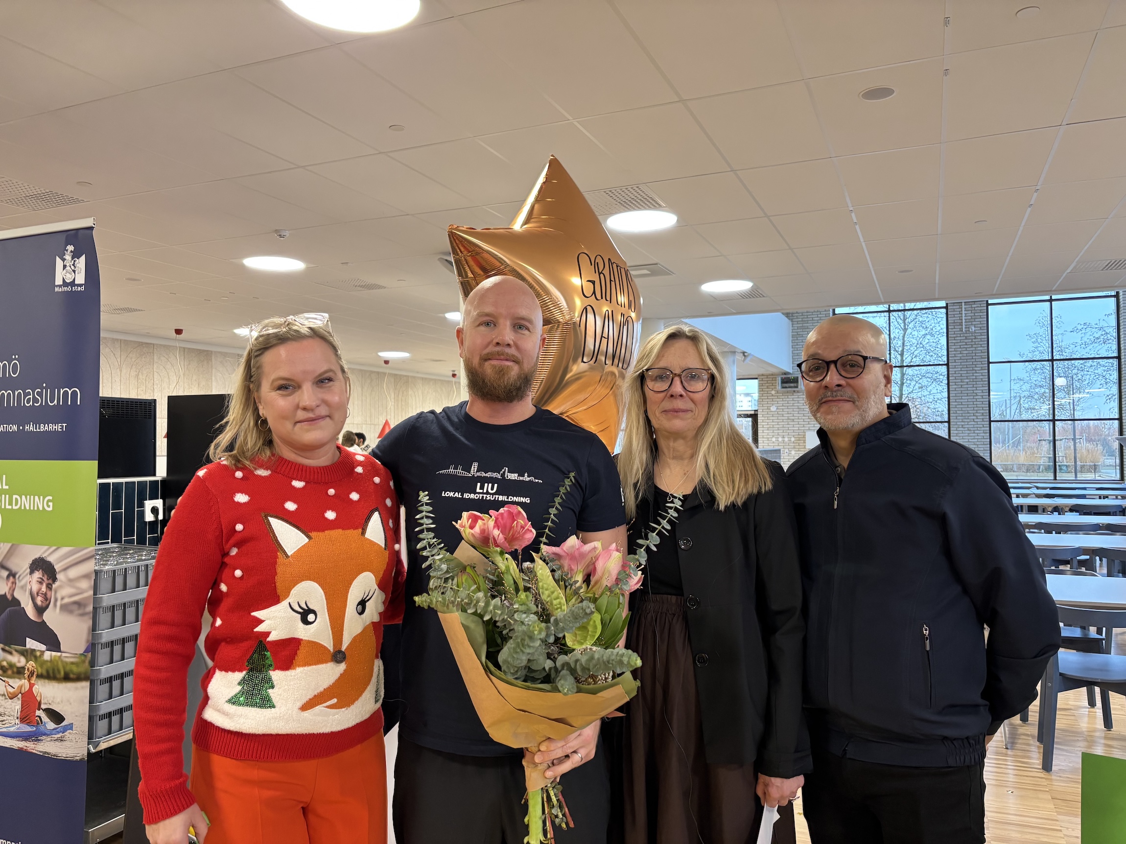 Fyra personer står i skolmatsal med blombukett och ballong med texten 'Grattis David' vid prisutdelning.